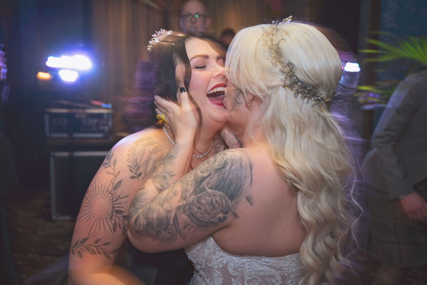 Two women share a joyful embrace on the wedding dance floor, one with tattooed arms.