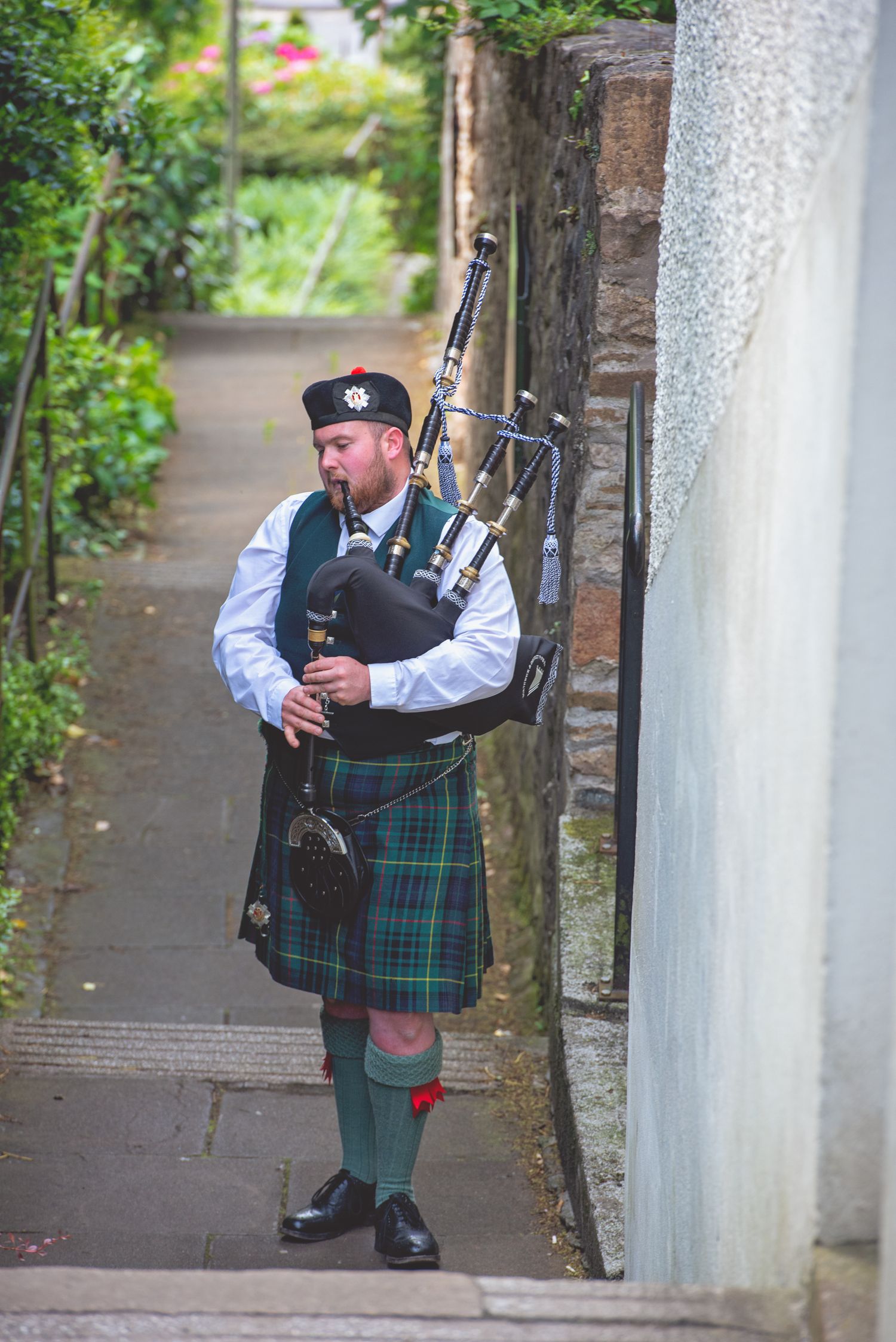 A bagpiper in traditional Scottish dress plays music in an outdoor stone corridor.