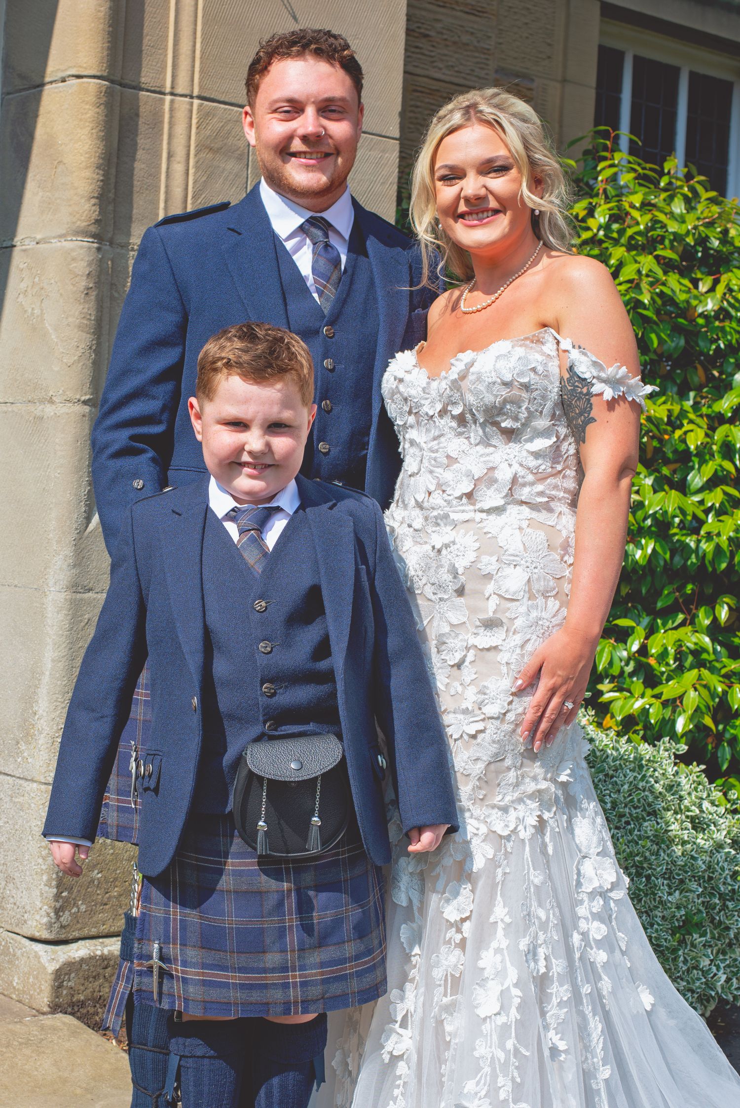Wedding party poses outdoors in navy suits and white lace dress against stone building.