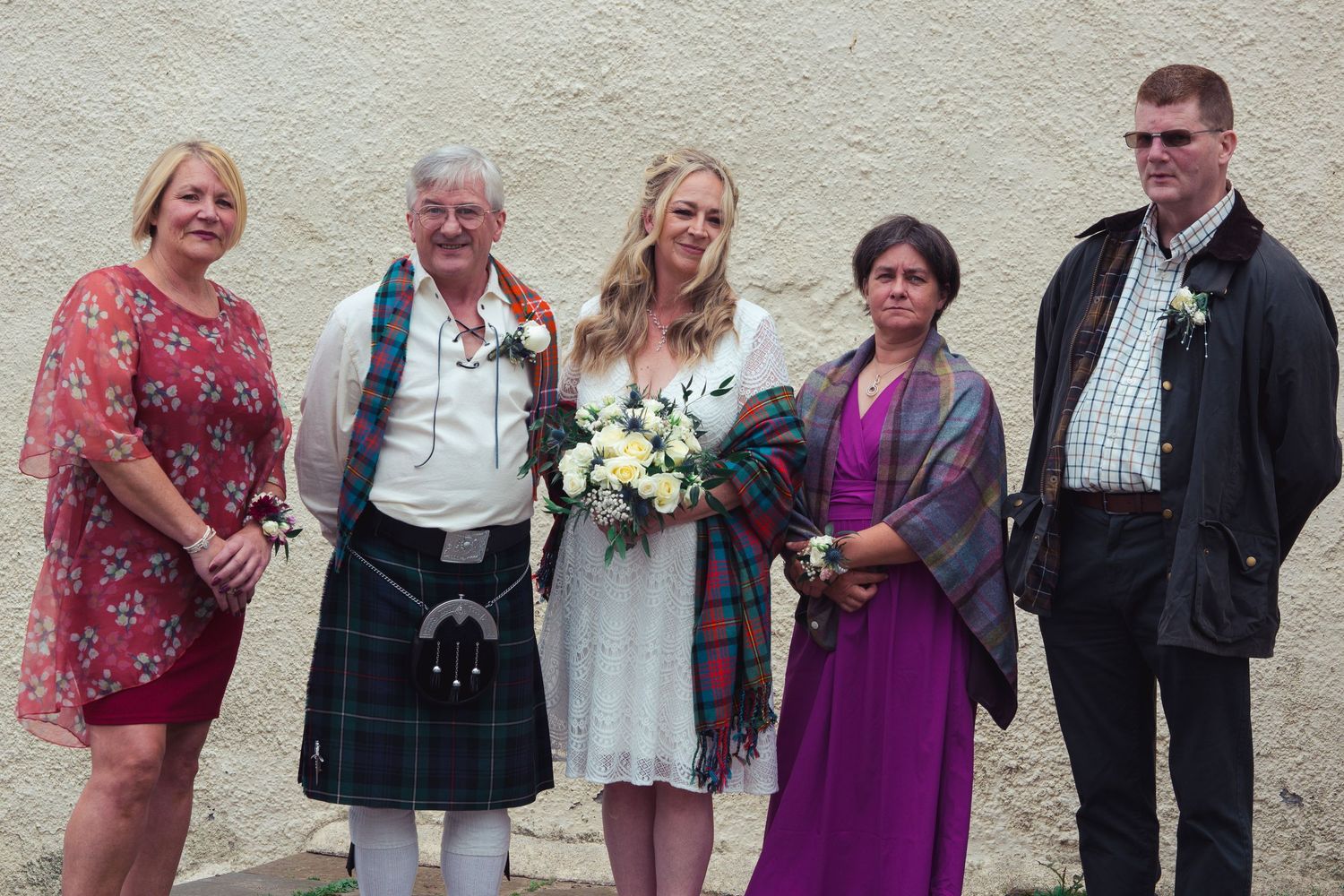 Wedding group photo with bride in white dress holding bouquet, man in Scottish kilt, and three guests posing outdoors.