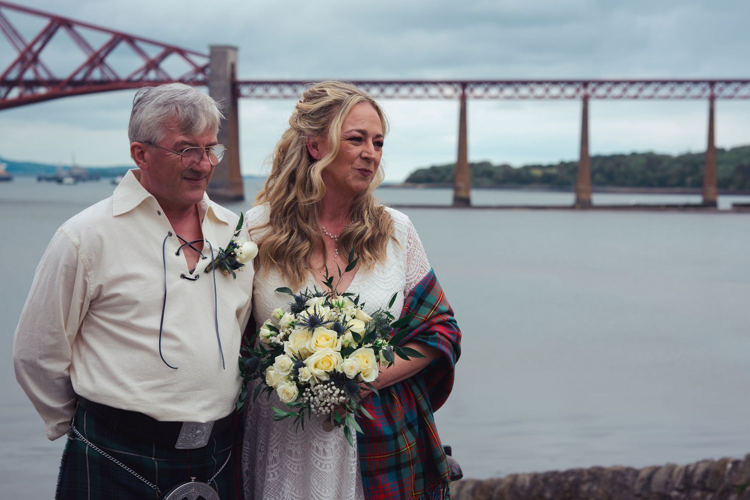 Bride with bouquet and man in kilt pose by the Forth Rail Bridge in Scotland on their wedding day.