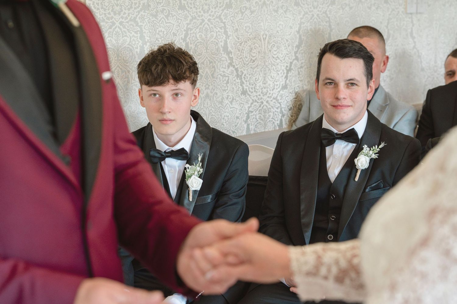 Two young men in black tuxedos with bow ties and boutonnieres watch a wedding ceremony with solemn expressions.