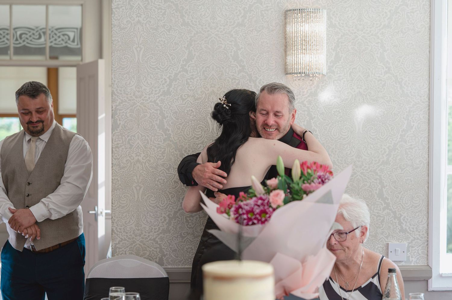 A bride hugs a smiling older man holding pink flowers at a wedding reception while guests look on.