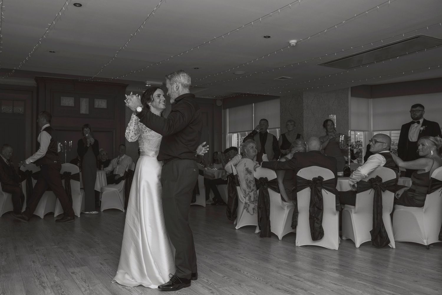Black and white photo of a couple sharing a first dance at a wedding reception with guests seated around the dance floor.