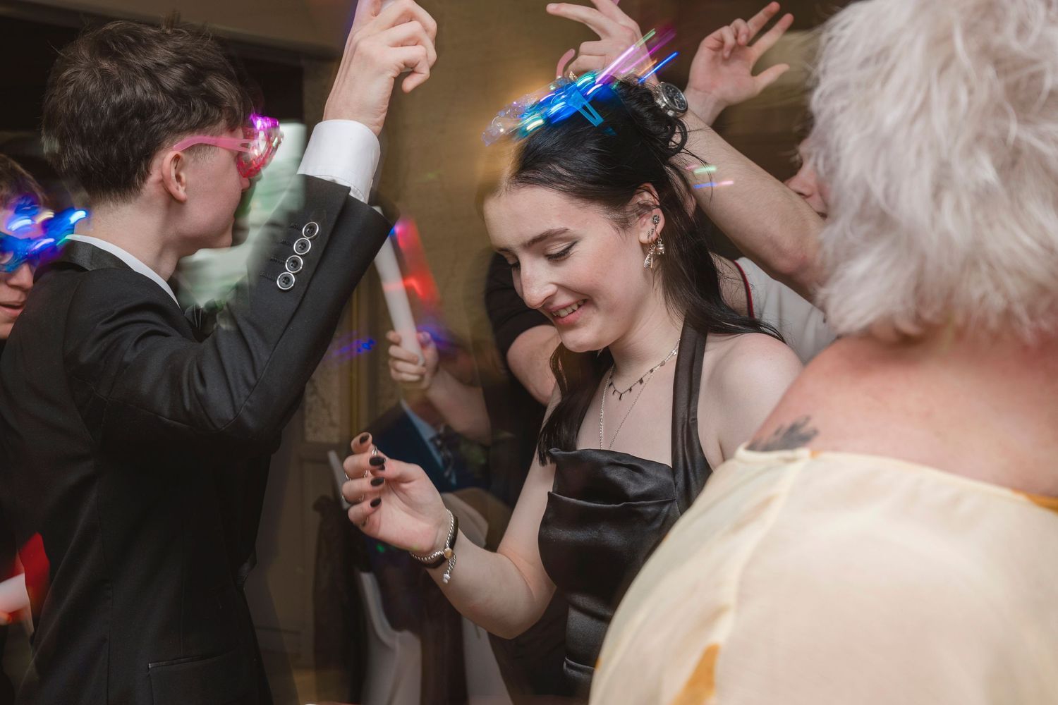 Young adults dancing at a party with colorful glow sticks, wearing formal attire at what appears to be a prom.