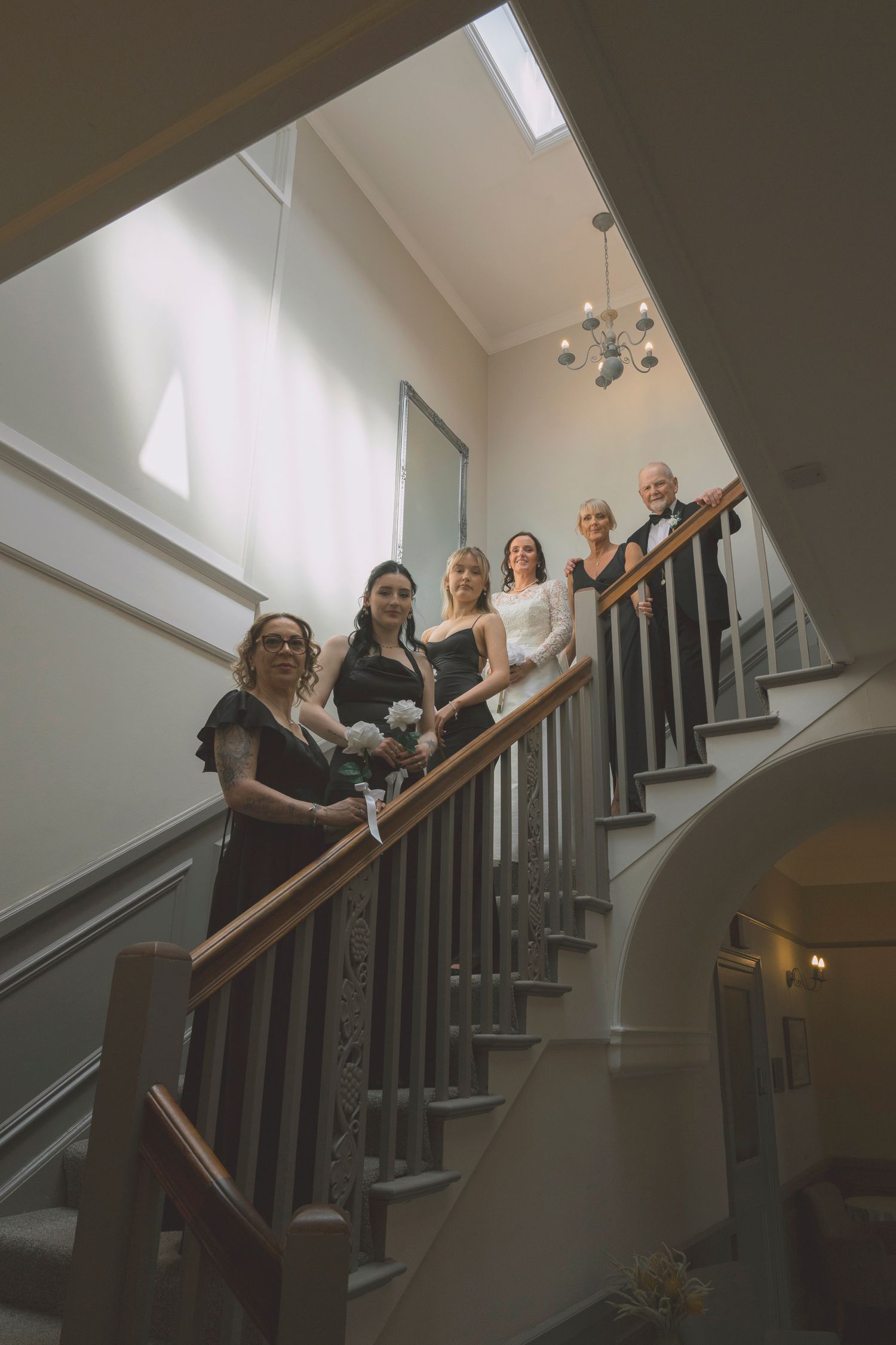 Wedding party posing on elegant staircase, bride in white gown with bridesmaids in black dresses and chandelier above.