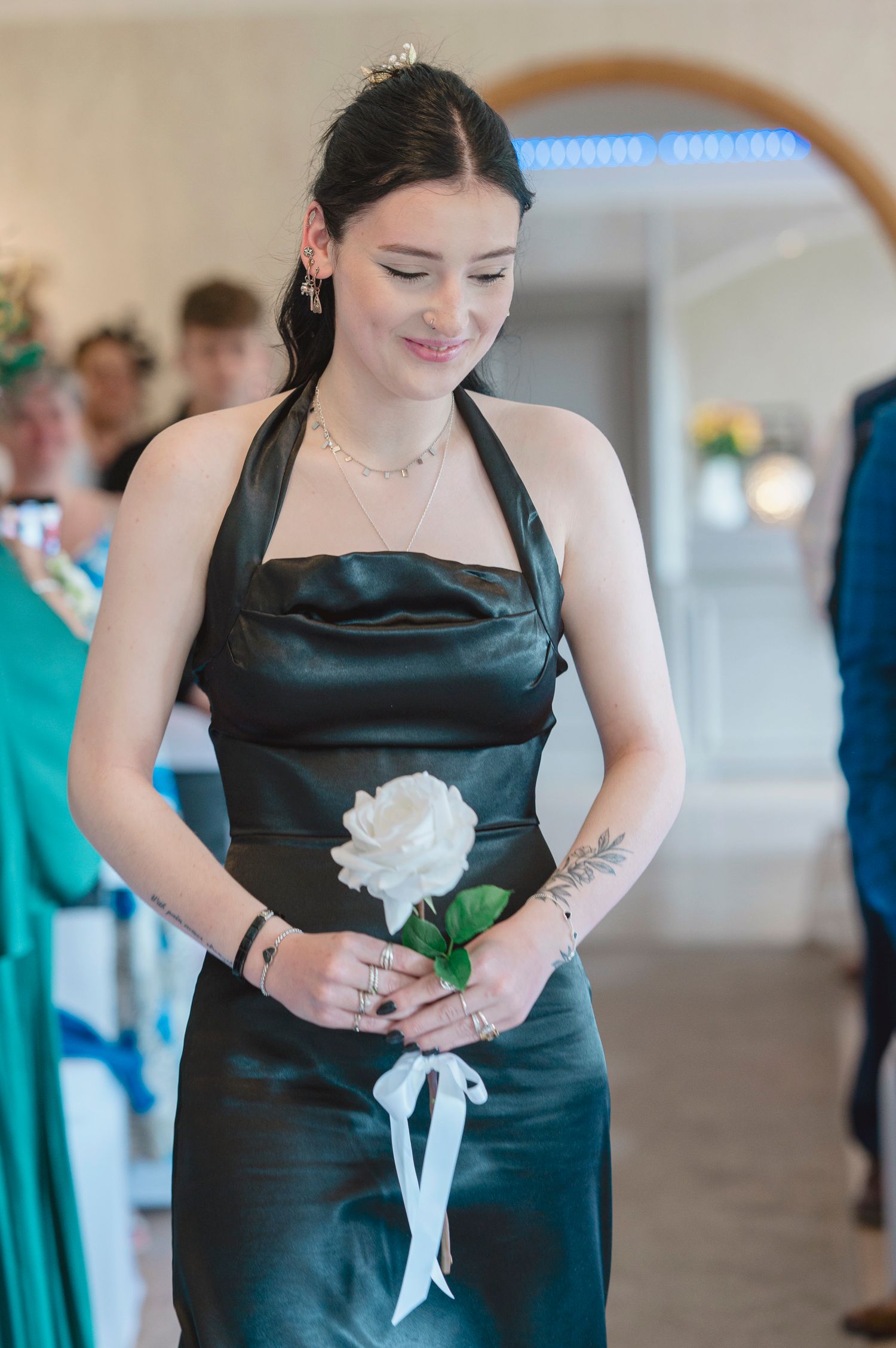 Young bridesmaid in black satin halter dress holding white rose with ribbon, smiling downward at indoor wedding ceremony.