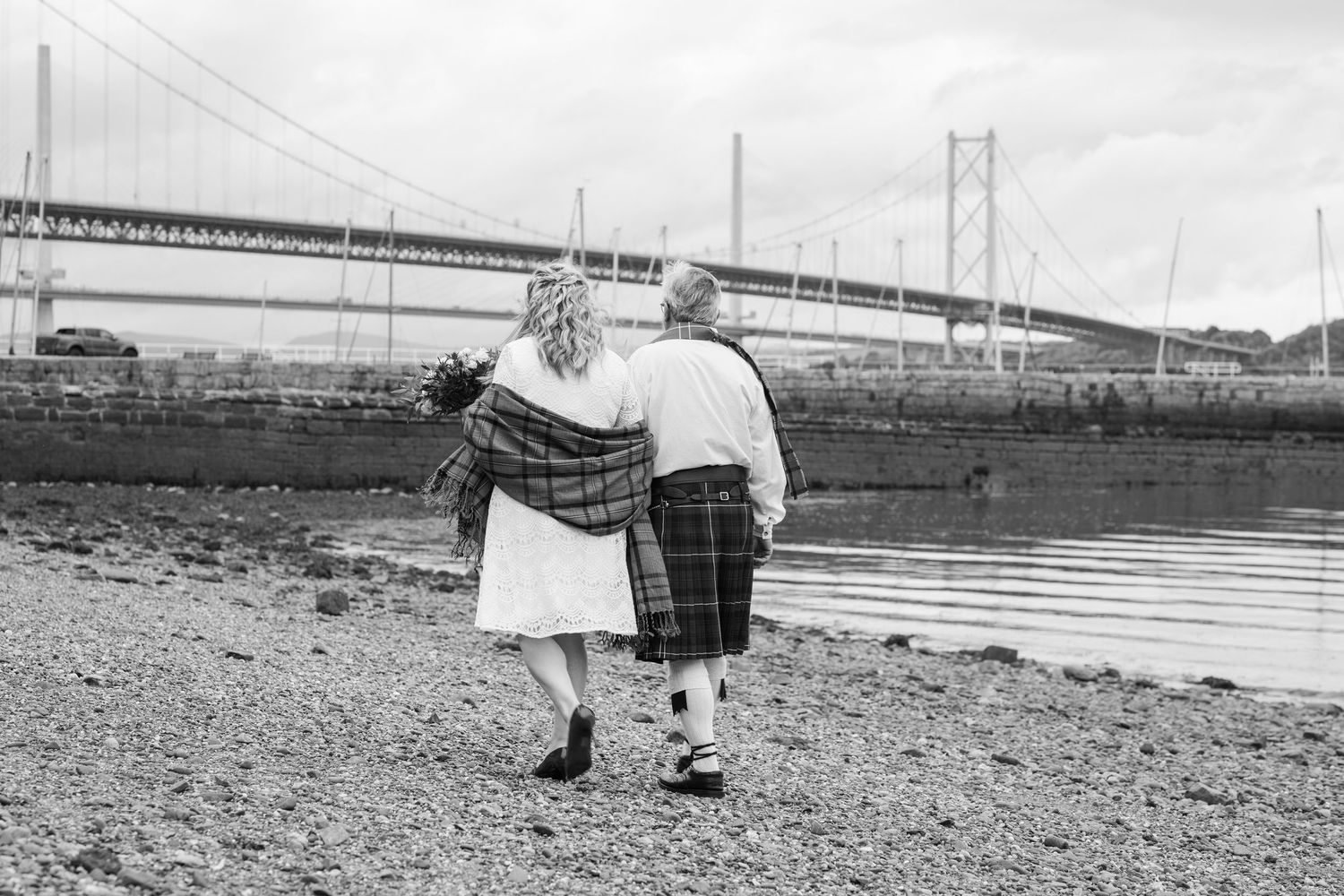 Black and white photo of a couple in Scottish attire walking along a rocky shore with the Forth Road Bridge in the background.