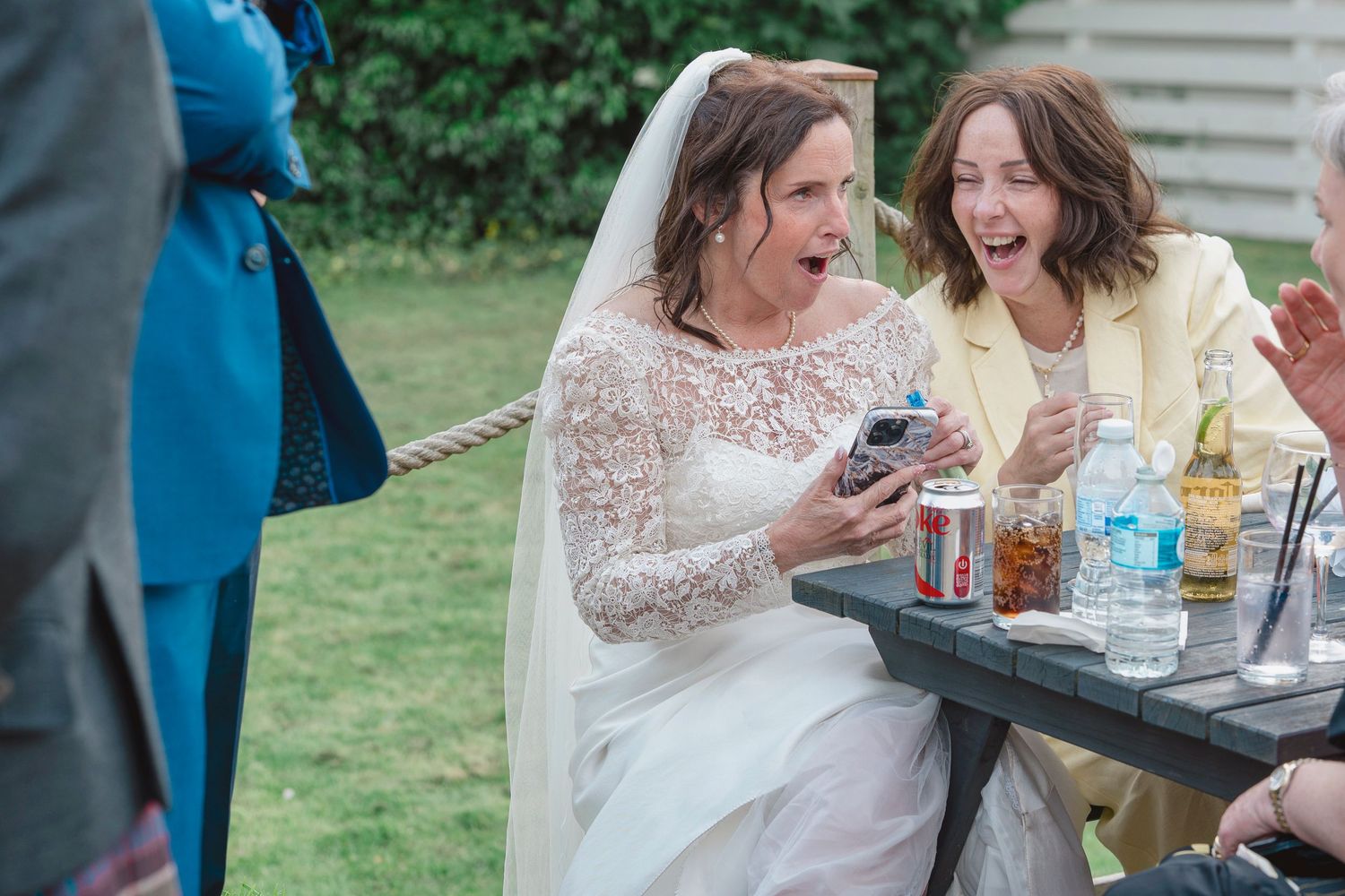 Bride in lace wedding dress laughing with female guest at outdoor wedding reception table with drinks.
