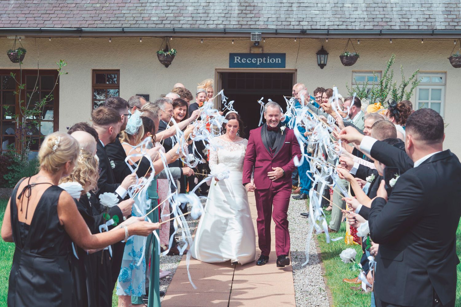 Bride and groom exit wedding reception venue through ribbon wand arch created by smiling guests outdoors.