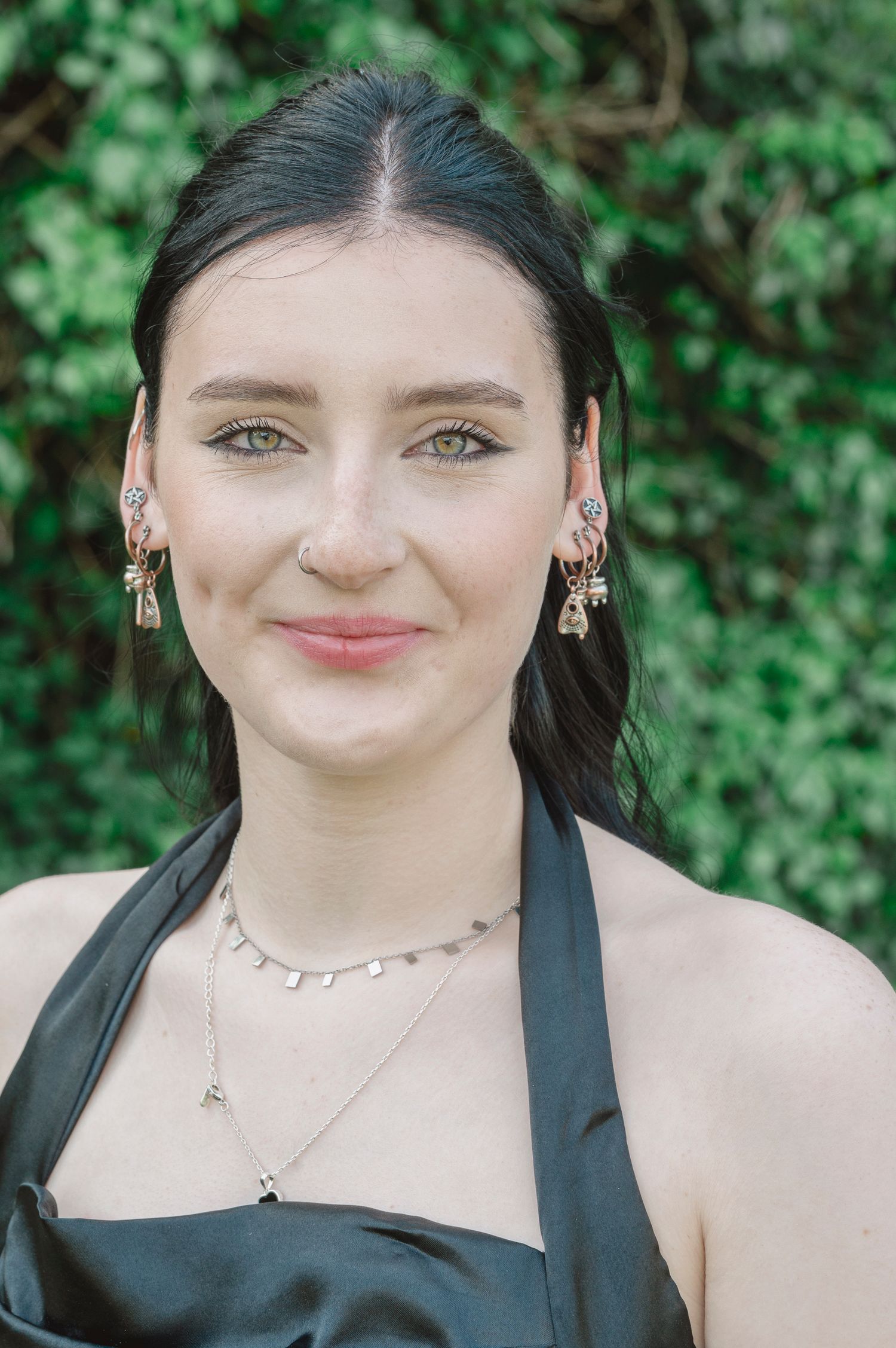 Young woman with dark hair wearing elegant earrings, septum ring, and layered necklaces in a black halter dress.