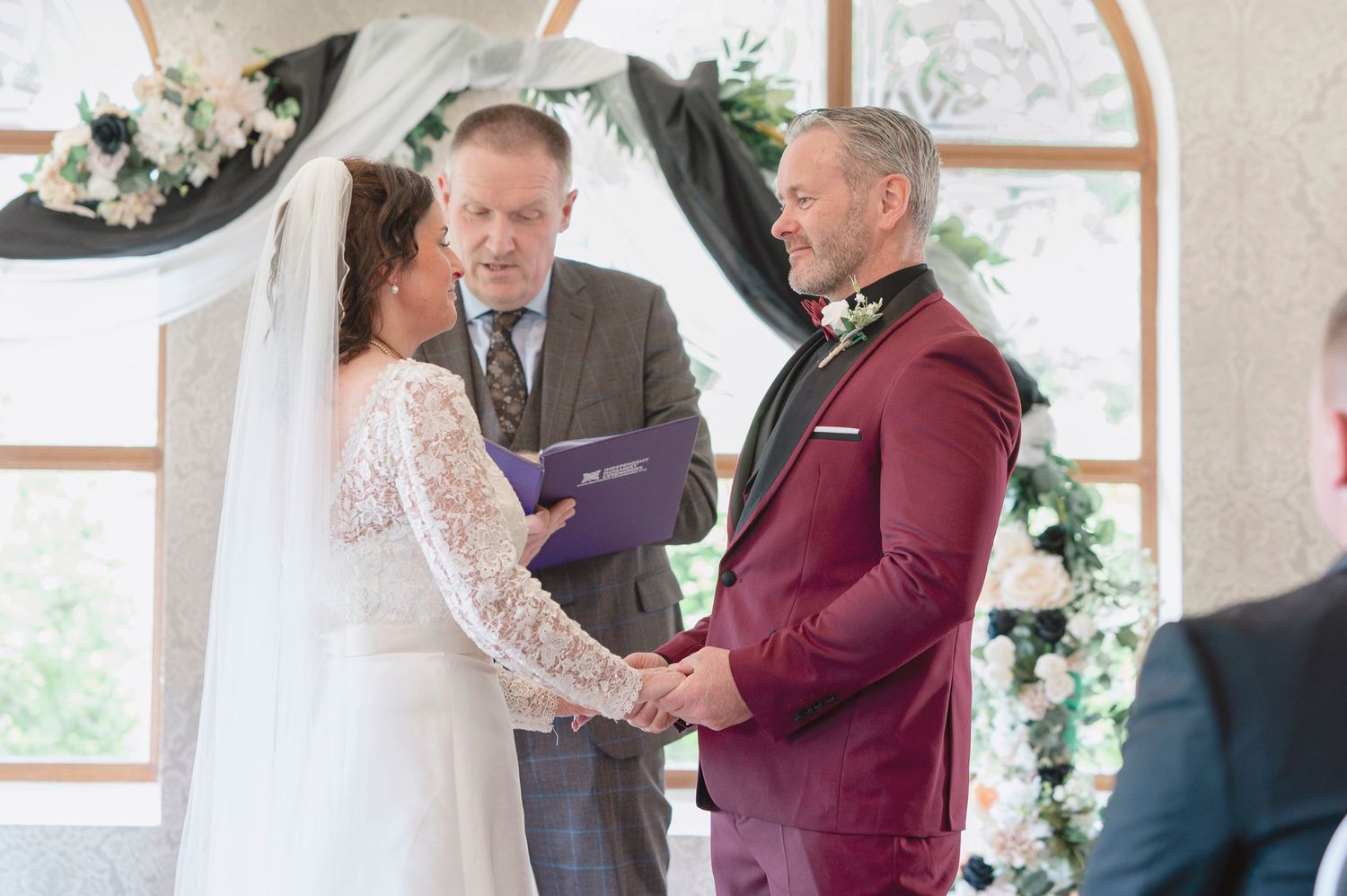 Bride and groom hold hands at wedding altar with officiant holding purple book during elegant indoor ceremony.