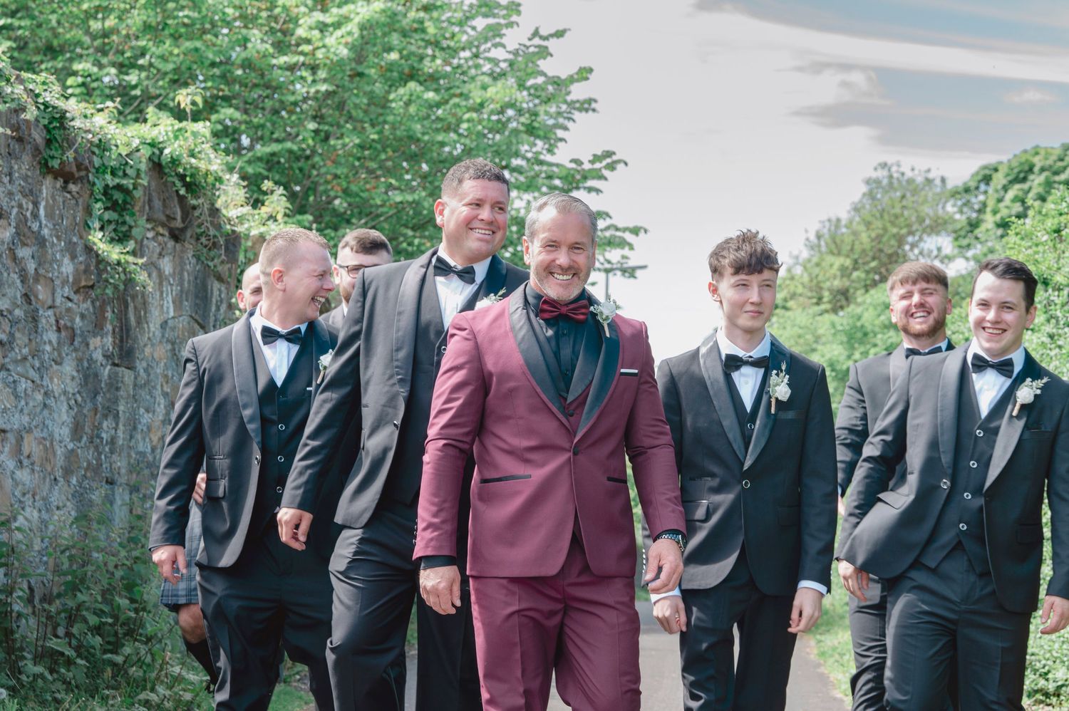 Groom in burgundy suit walks with groomsmen in dark suits and bow ties outdoors at a wedding.