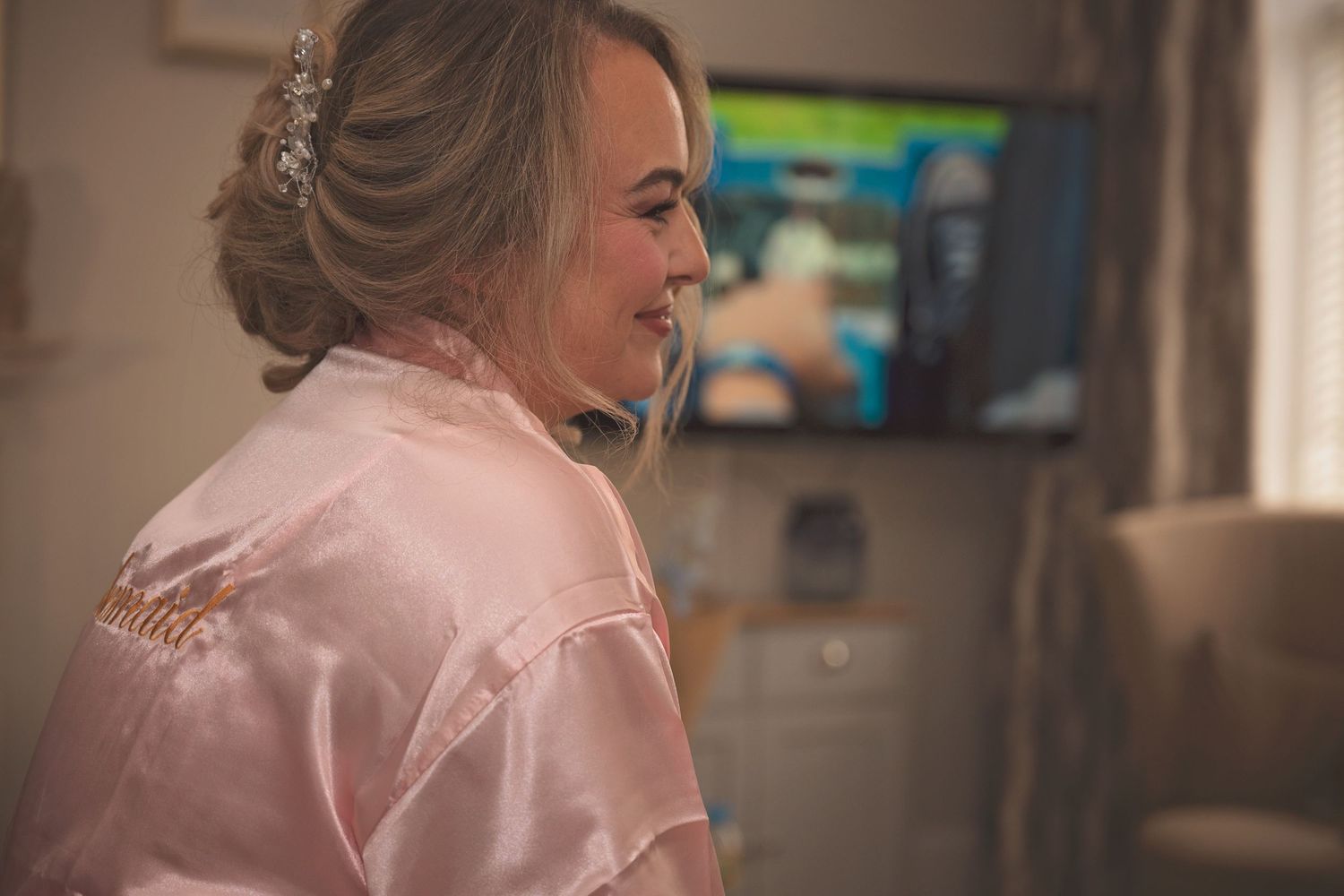 Bride in pink satin robe with elegant updo hairstyle and hair accessories during wedding morning preparations.