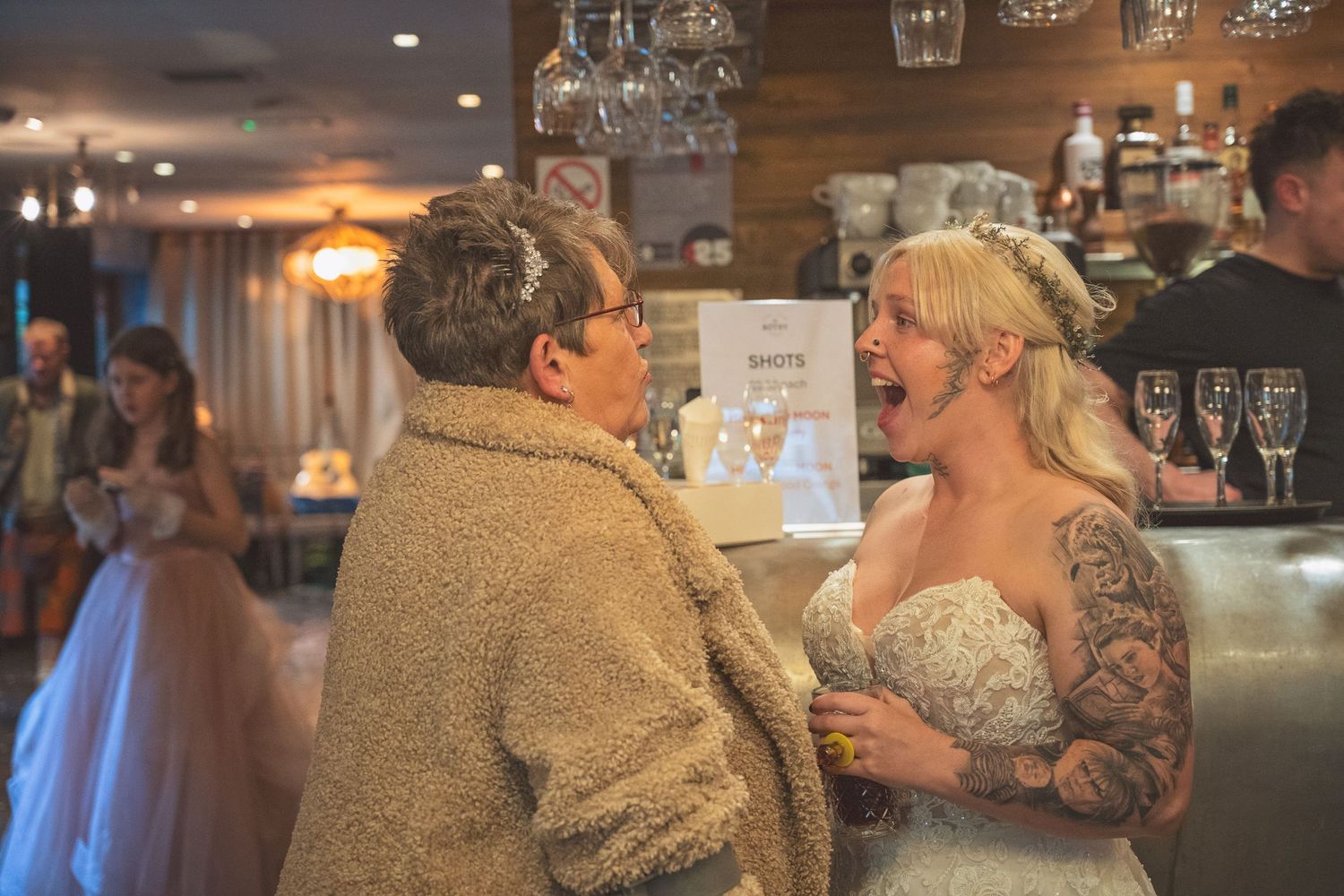 Tattooed bride in white lace gown laughing with older woman guest at wedding reception bar.