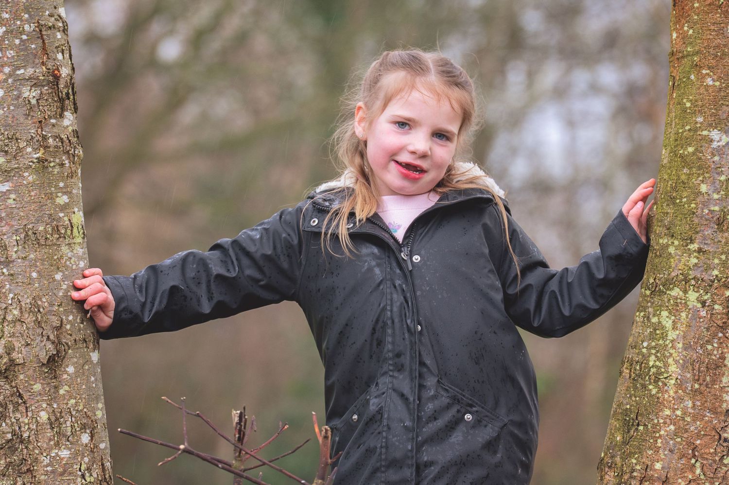 A child in a black jacket stands between two tree trunks with arms outstretched on a cloudy day.