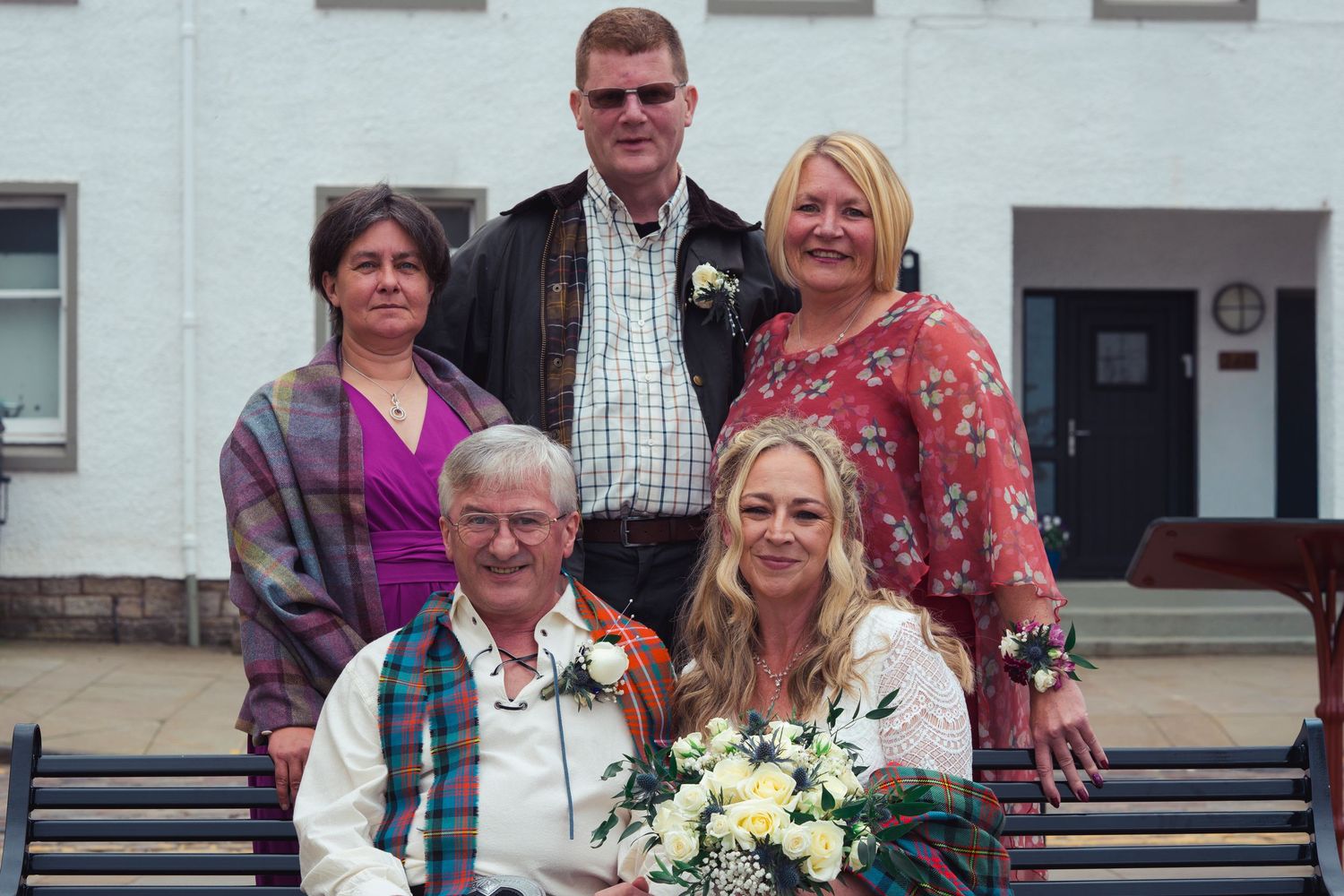 A family group of five poses for a wedding photo, with a couple seated on a bench holding a bouquet.