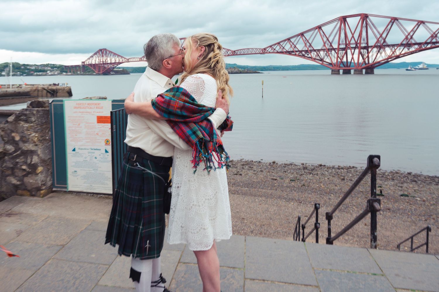 Couple kissing in kilts near the Forth Bridge in Scotland on a cloudy day.