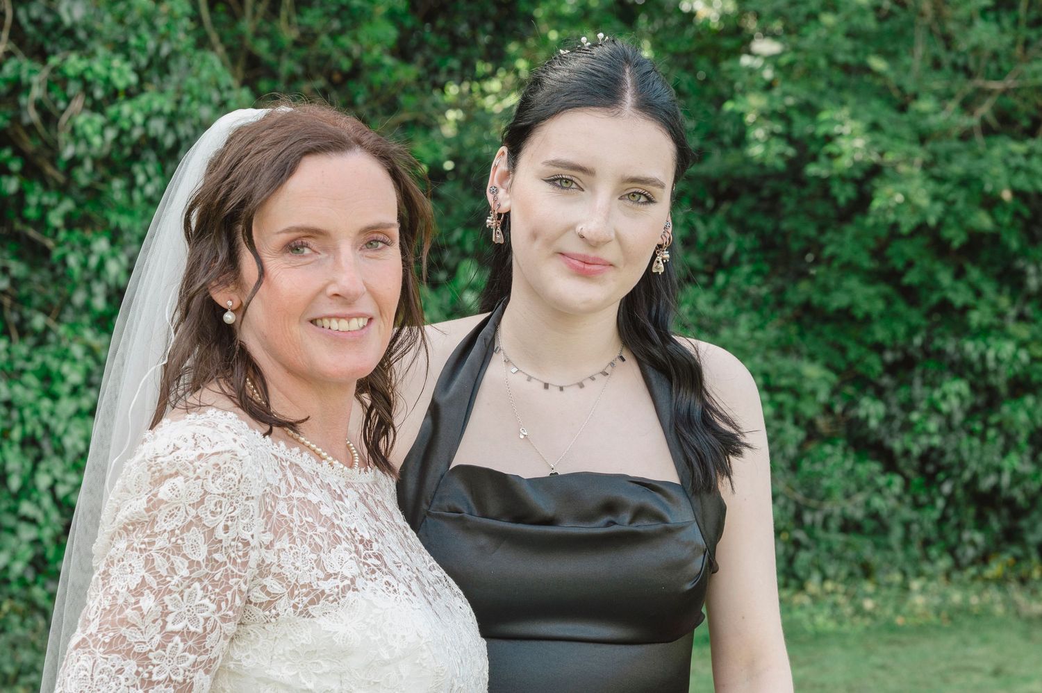 Bride in lace veil and woman in black halter dress pose together outdoors at a wedding celebration.