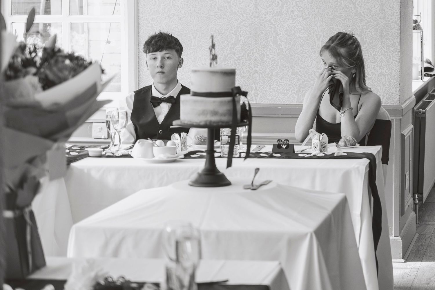 Black and white photo of two women sitting at a wedding reception table with a tiered cake, one wiping tears.