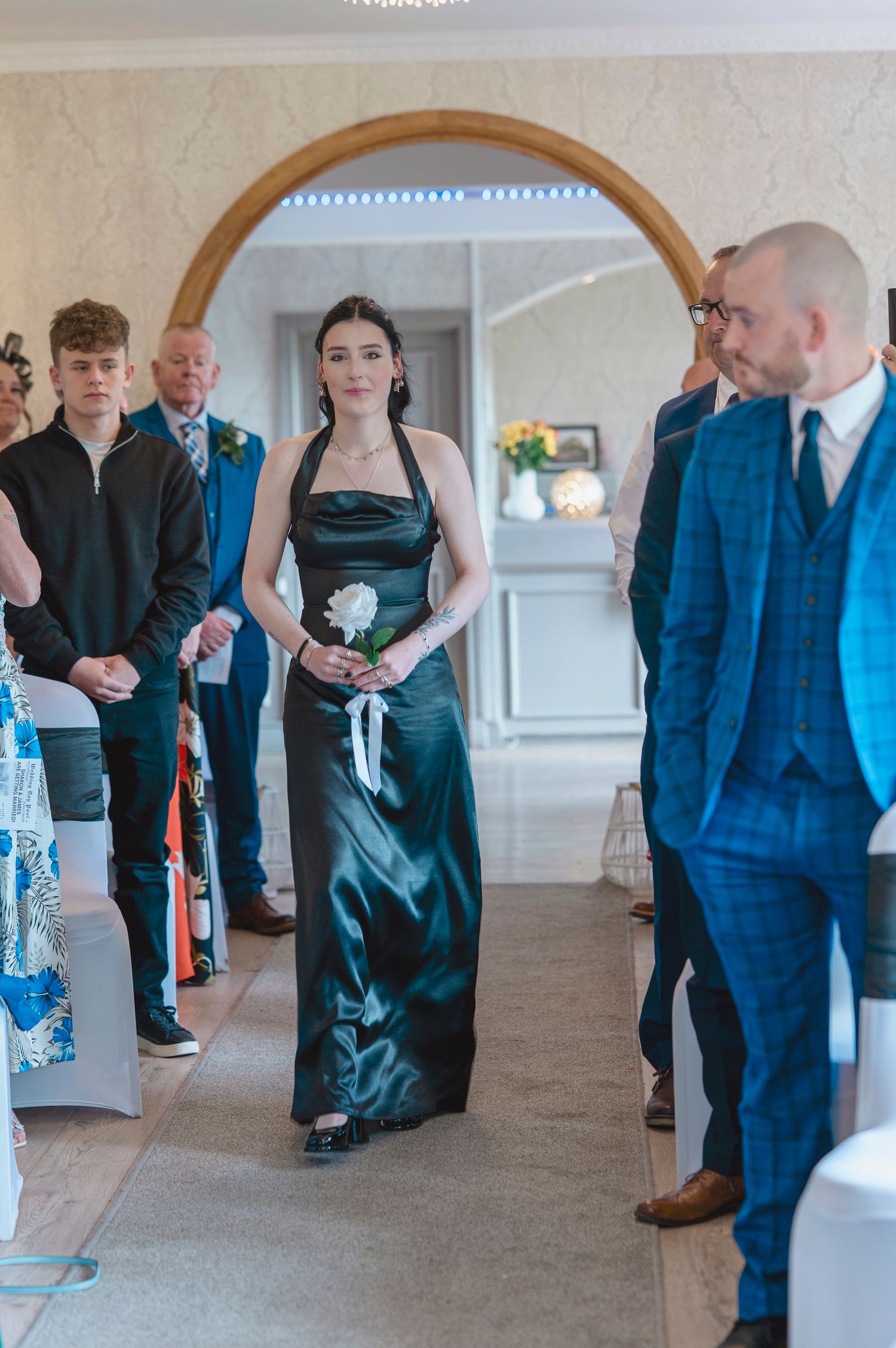Bridesmaid in black satin halter gown walks down wedding aisle holding white flower, guests seated on either side.