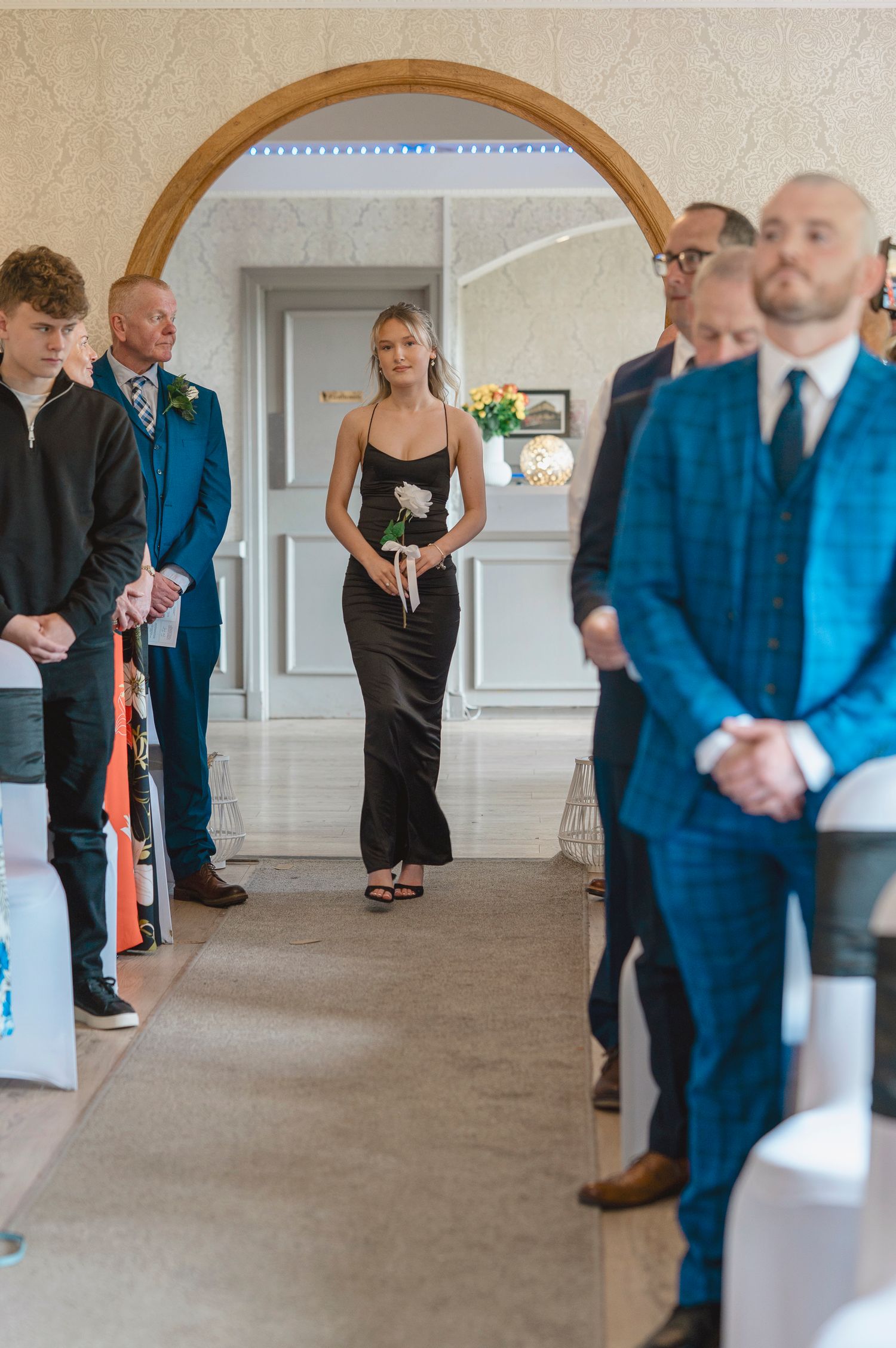 A bridesmaid in a black dress walks down the aisle holding a white rose at an elegant wedding ceremony.