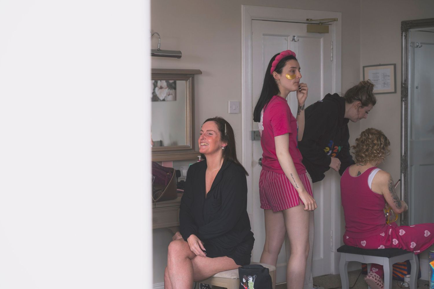 Women getting ready in a room, one seated smiling, another standing in pink dress with headband.