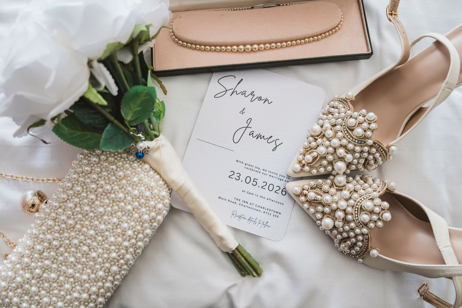 Wedding flat lay with pearl heels, bouquet, invitation for Sharon & James dated 23.05.2020, pearl clutch, and nude accessories.