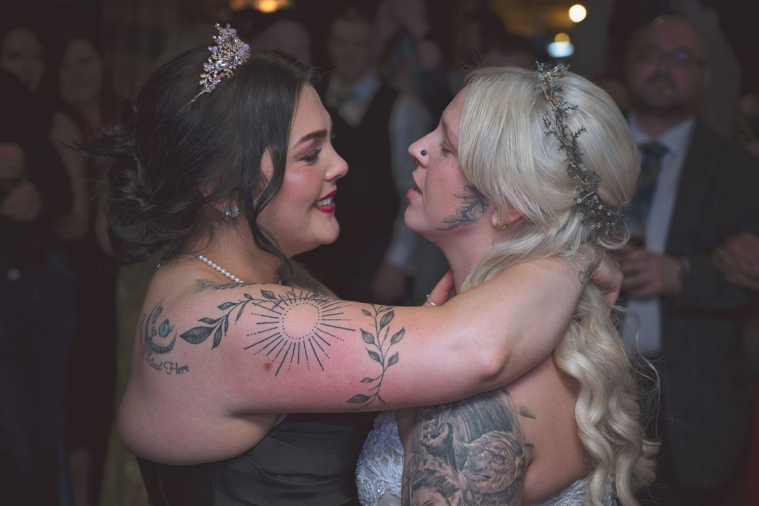 Two brides in wedding attire with floral tattoos share a tender dance moment at their wedding reception.