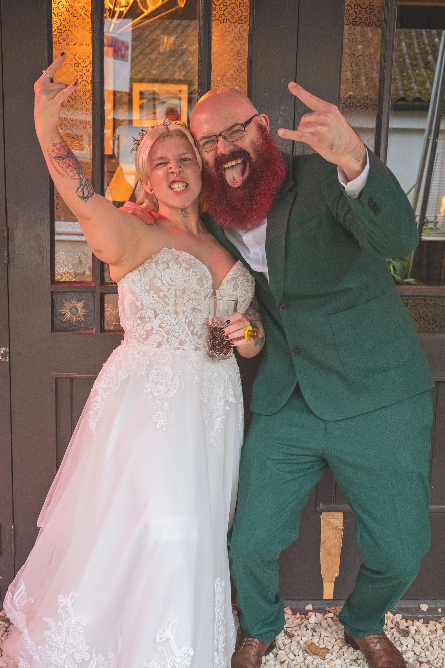 Newlywed couple celebrate joyfully outside venue, bride in lace gown and groom in green suit with red beard.