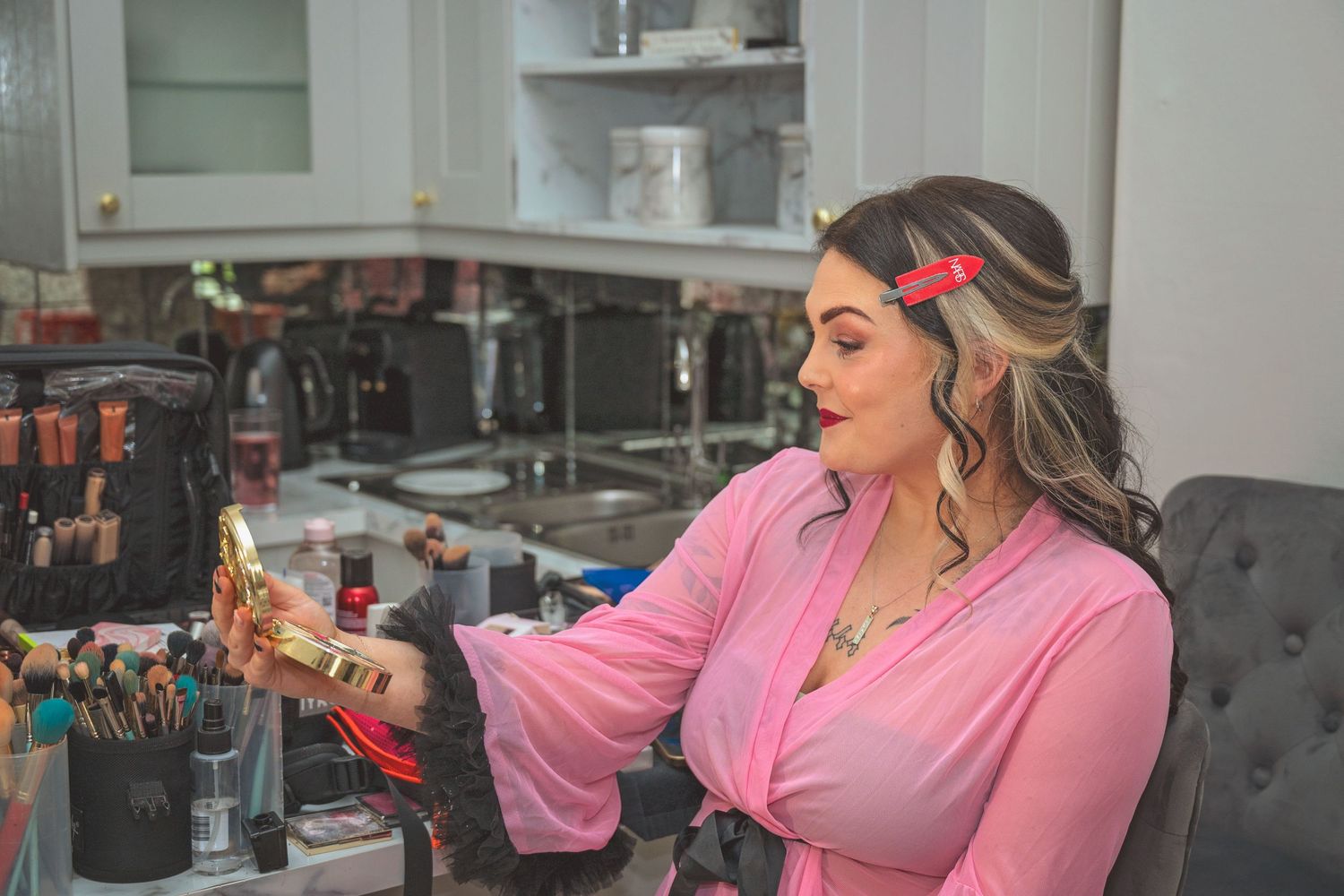Woman in pink blouse examines gold makeup product at vanity with brushes and beauty tools.