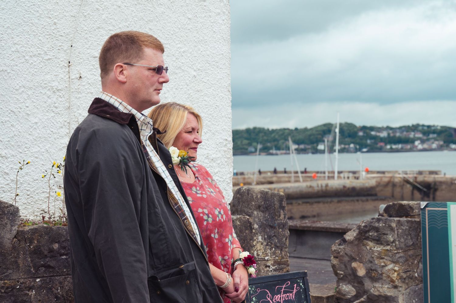 Couple standing together by a harbor wall, man in dark jacket, woman in floral dress, cloudy coastal backdrop.