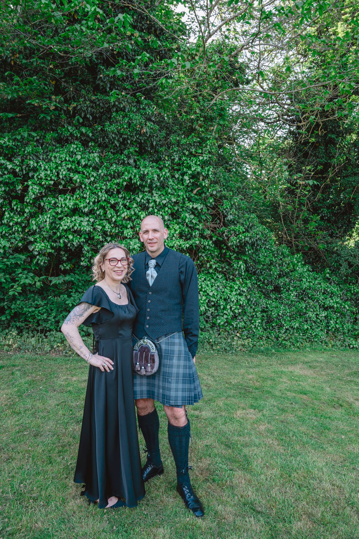 Couple posing outdoors in formal attire, woman in black gown and man in traditional Scottish kilt with sporran.