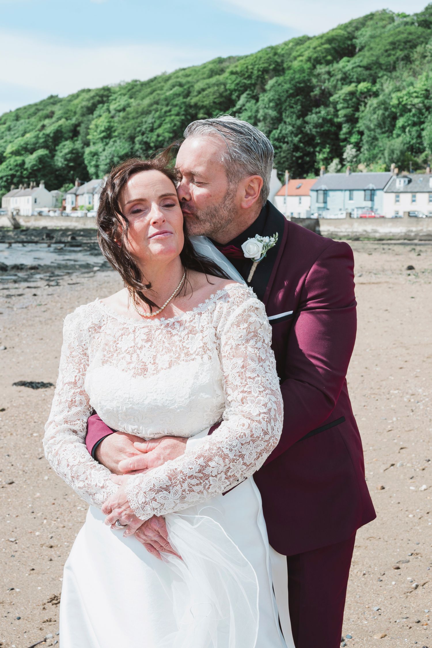 A groom in a burgundy suit kisses his bride in a lace wedding dress on a sandy beach.