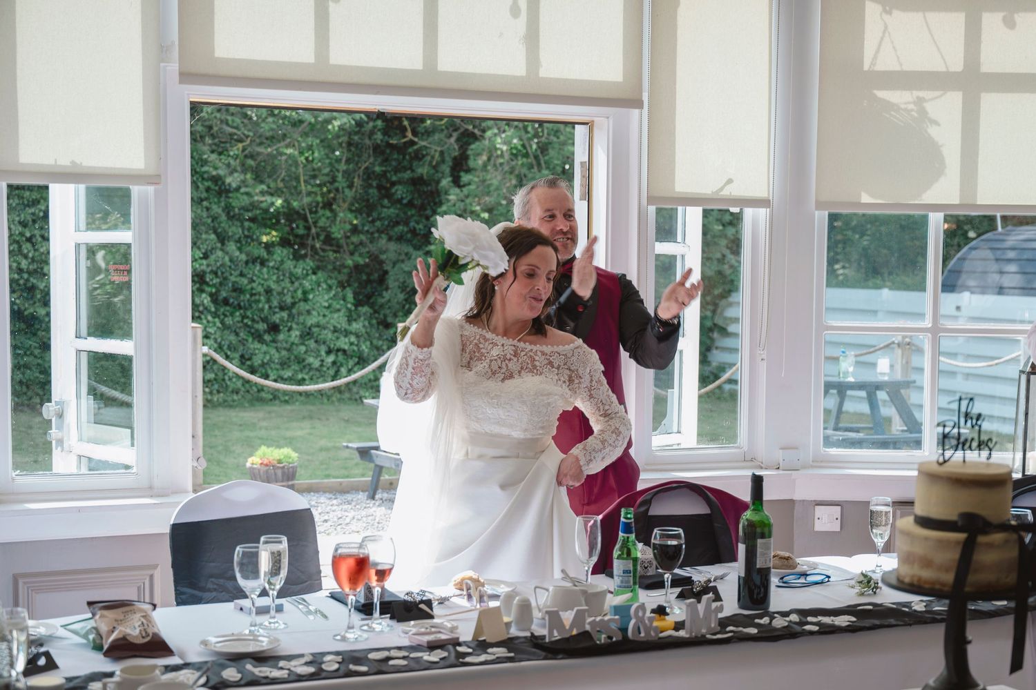 Bride and groom celebrating joyfully at wedding reception table with drinks and decorations near large windows.