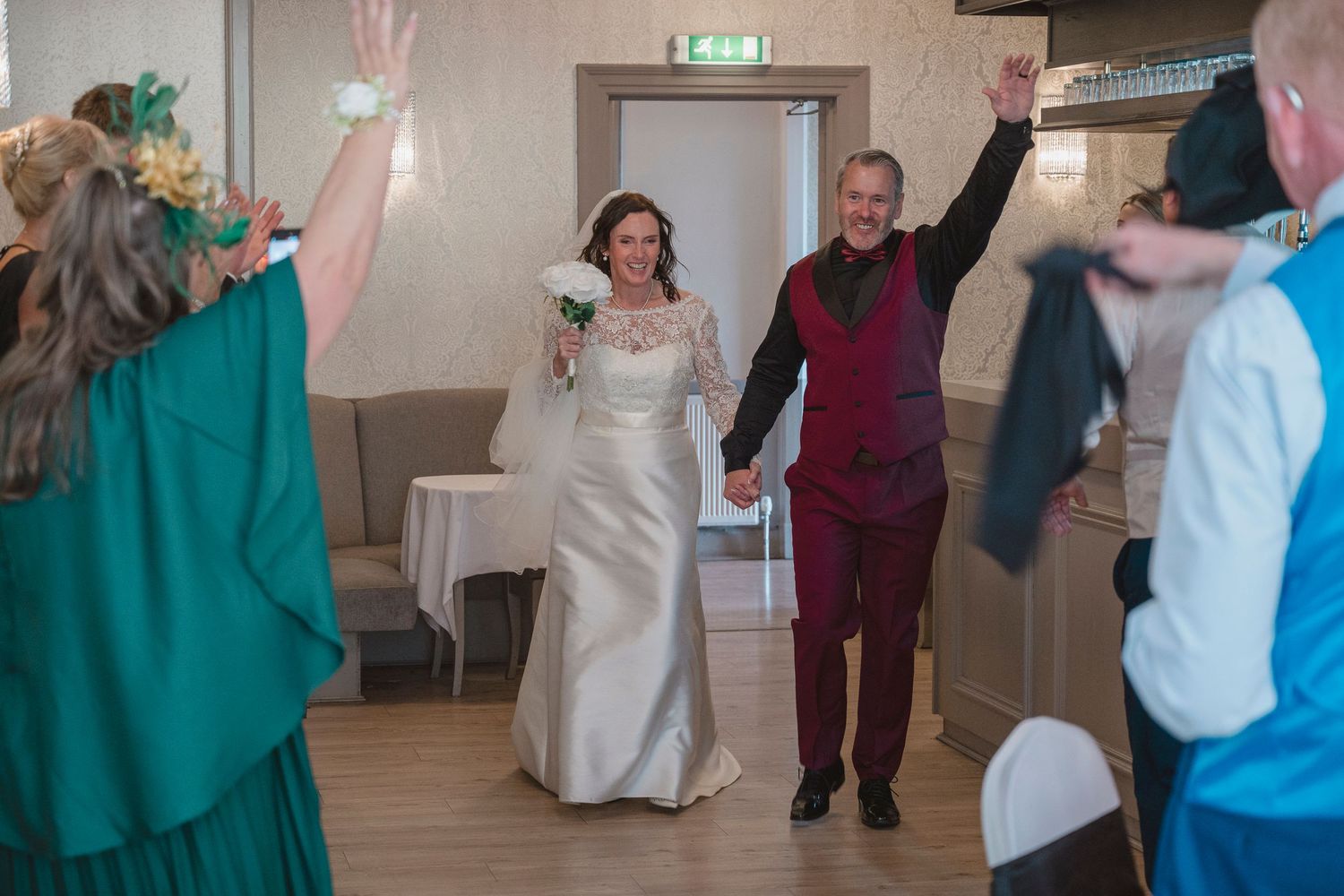 Bride and groom enter wedding reception hall holding hands, guests cheering with raised arms in celebration.