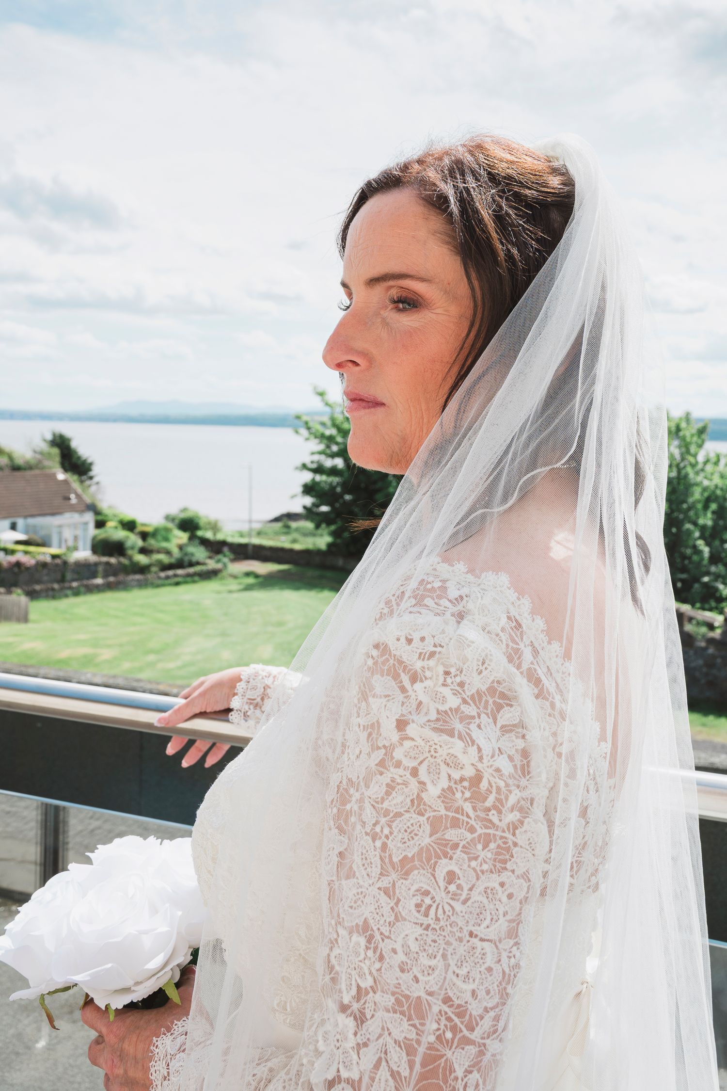 Bride in lace long-sleeve gown and veil holds white peony bouquet on balcony overlooking scenic waterfront.