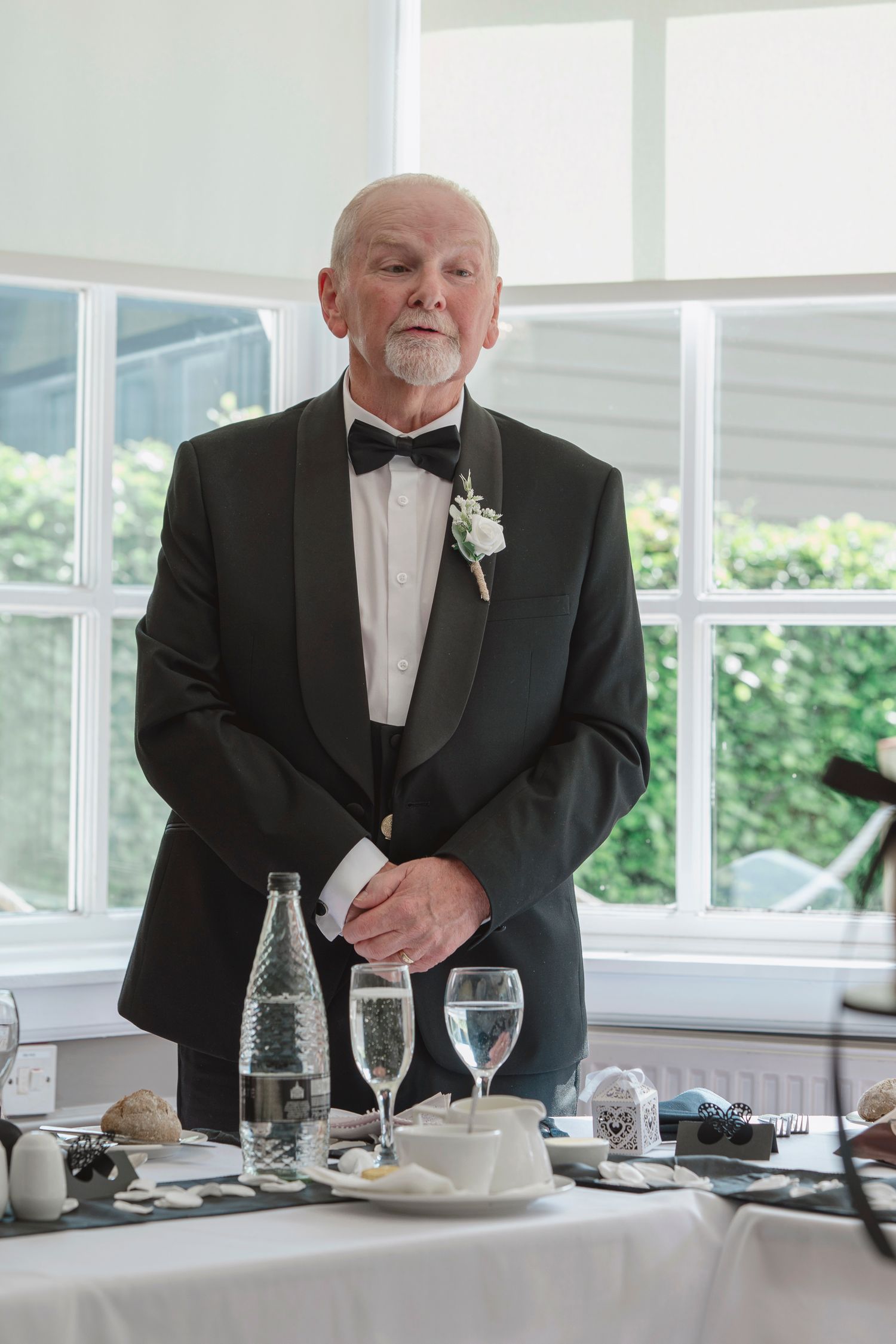 Elderly man in black tuxedo with boutonniere stands at wedding reception table with champagne glasses.