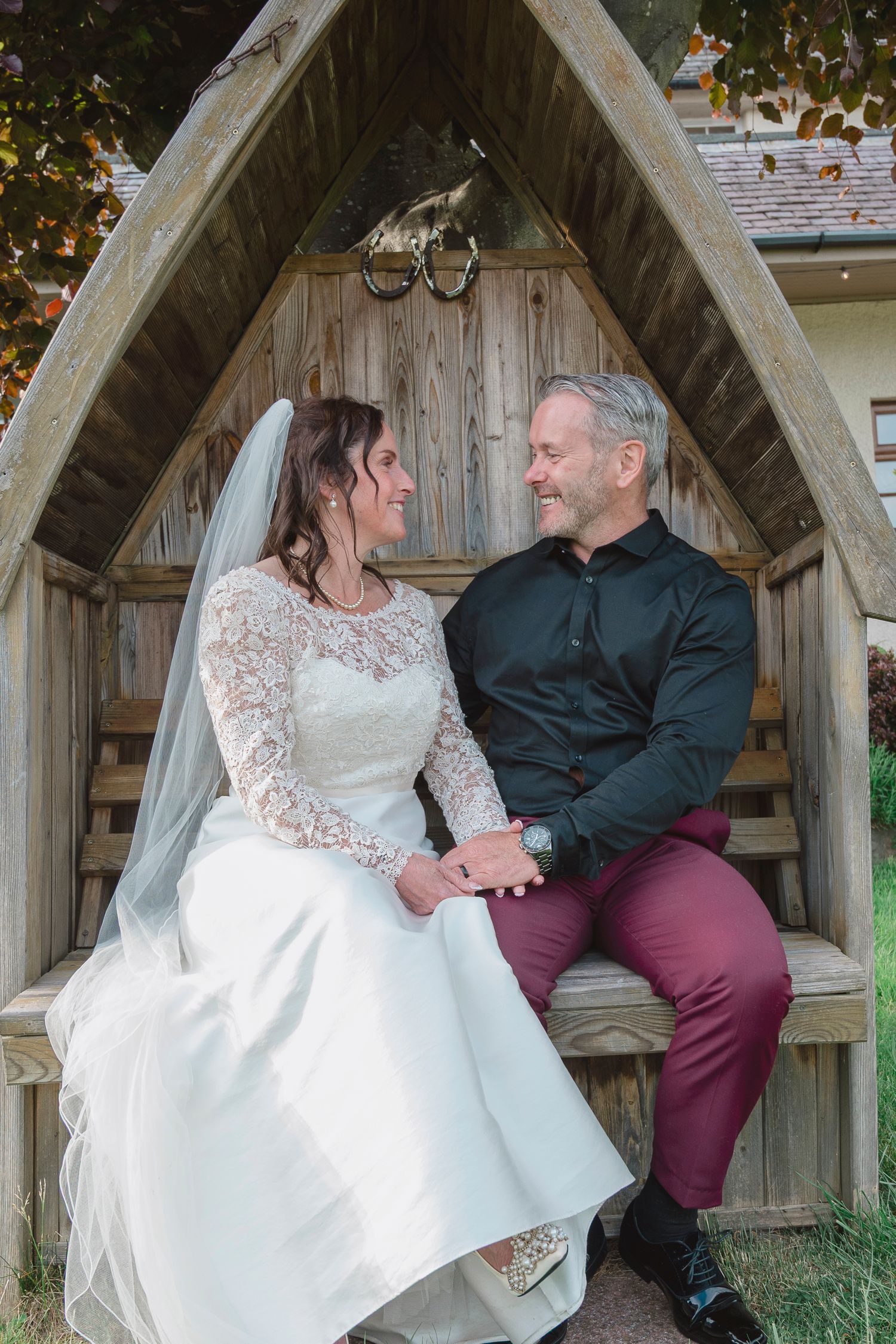 A bride and groom sit together in a wooden arbor, holding hands and smiling at each other on their wedding day.
