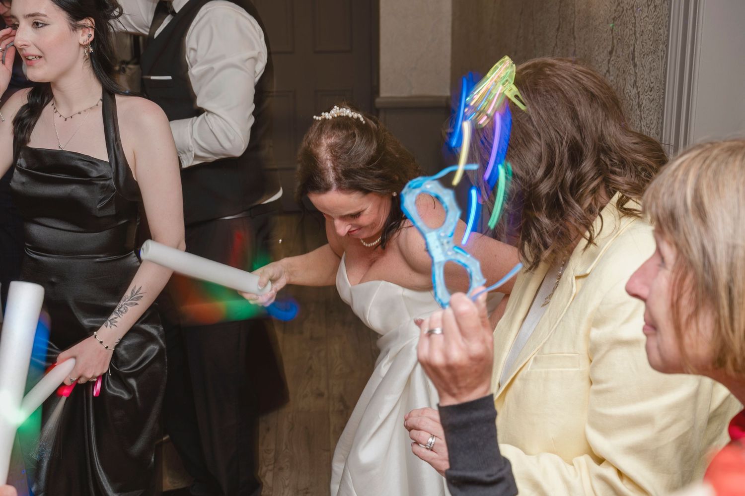 Bride in white dress dancing with guests holding glow sticks and balloon animals at a lively wedding reception.