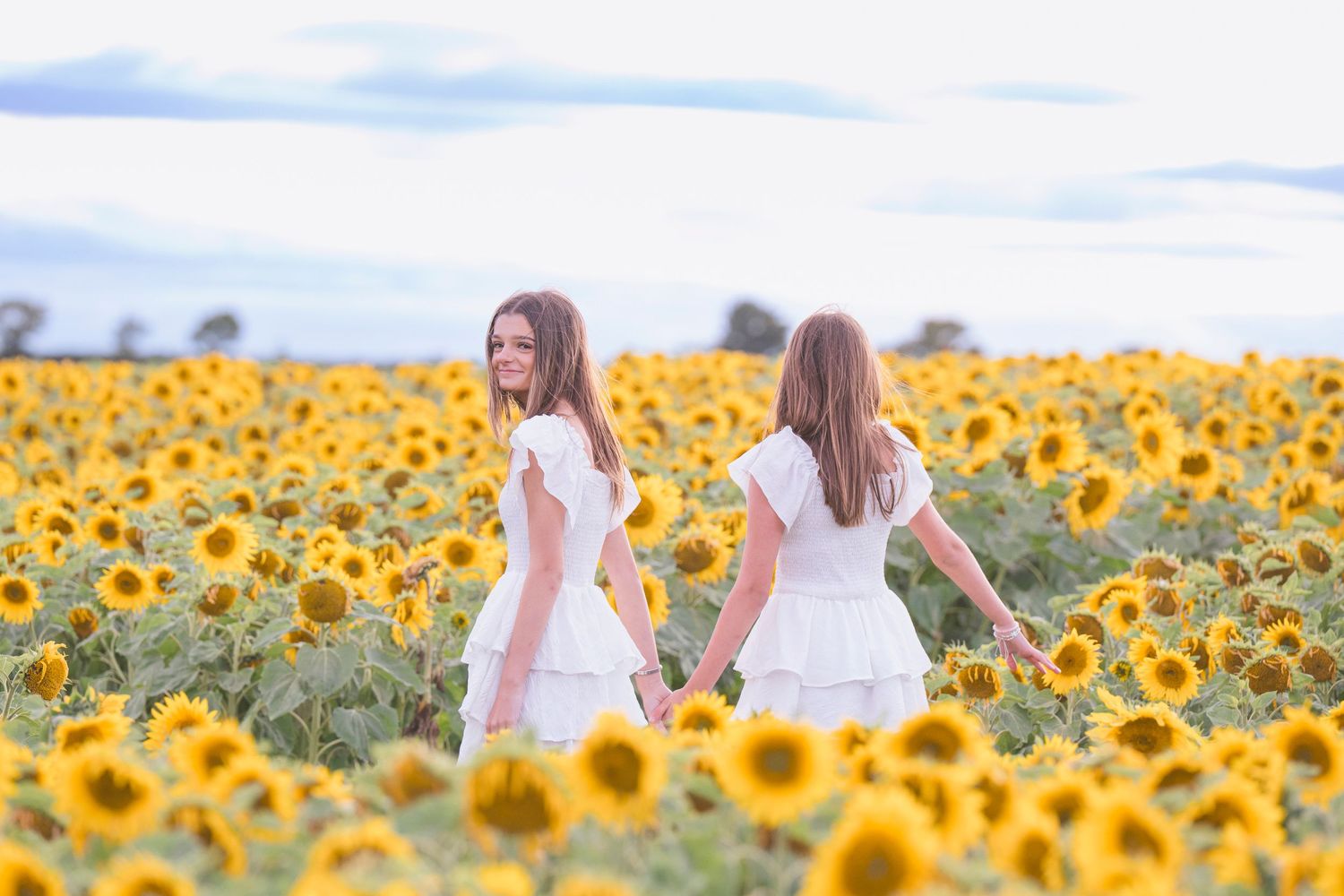 Two girls in white dresses hold hands in a vast sunflower field under a cloudy sky.