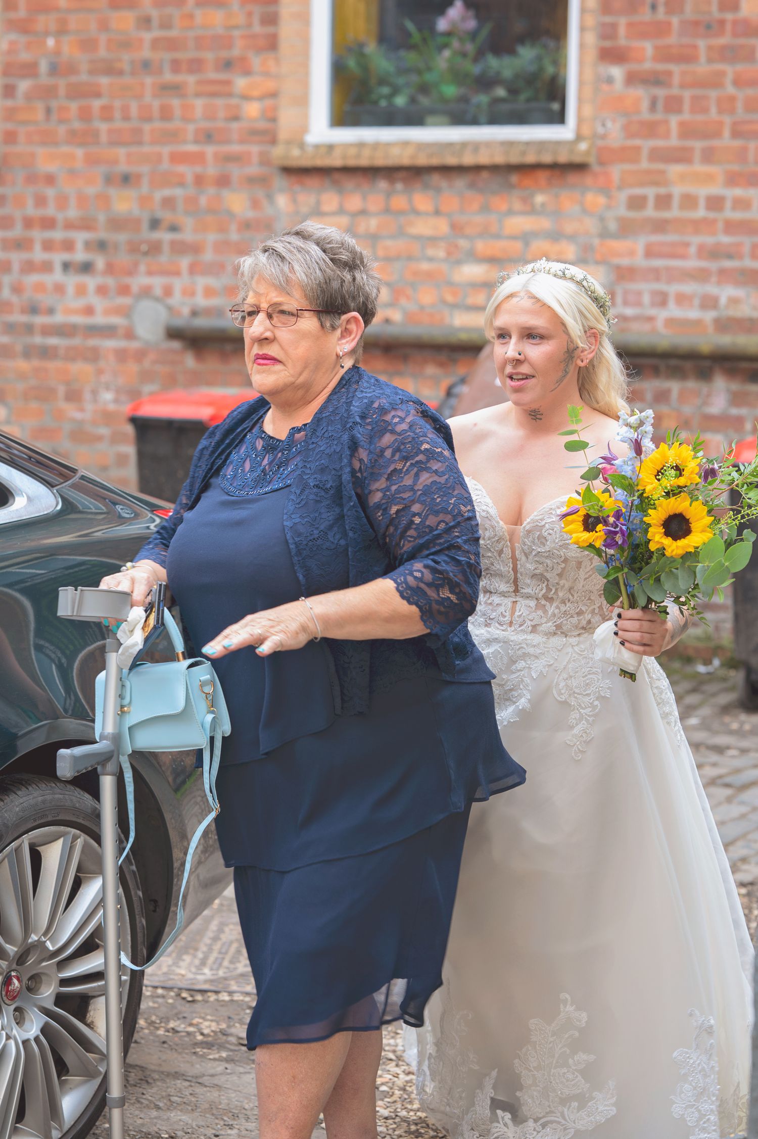 Bride in lace wedding gown holding sunflower bouquet walks with older woman in navy dress outside brick building.