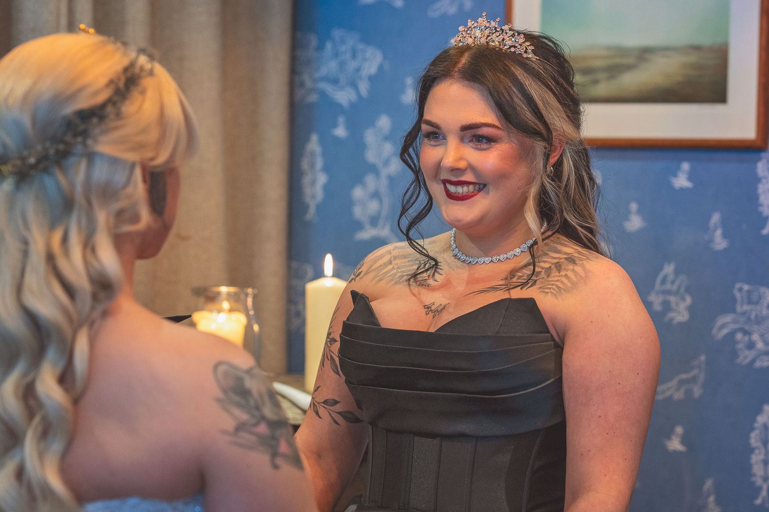 A tattooed woman in a black strapless gown and tiara smiles during a wedding ceremony.