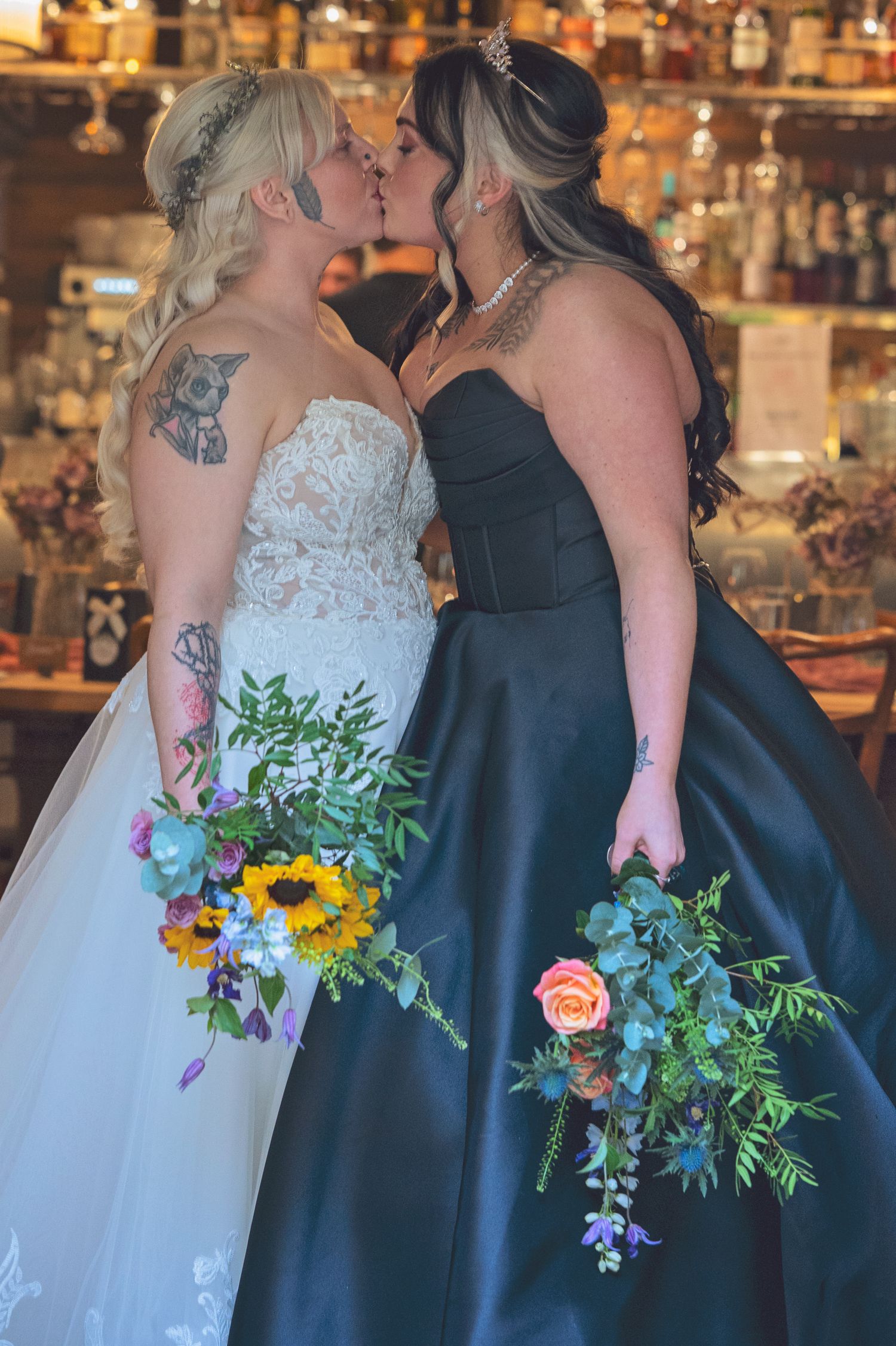 Two brides share a kiss, one in white lace gown and one in black dress, holding colorful bouquets.