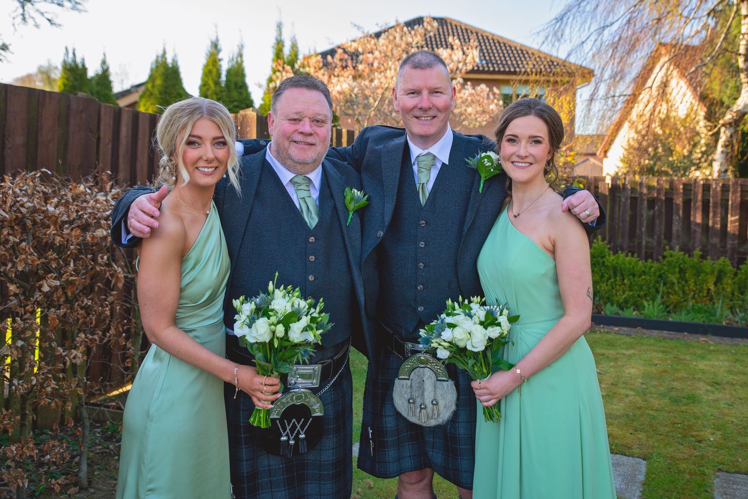 Wedding party members in mint green dresses and dark suits pose together outdoors in a garden setting.