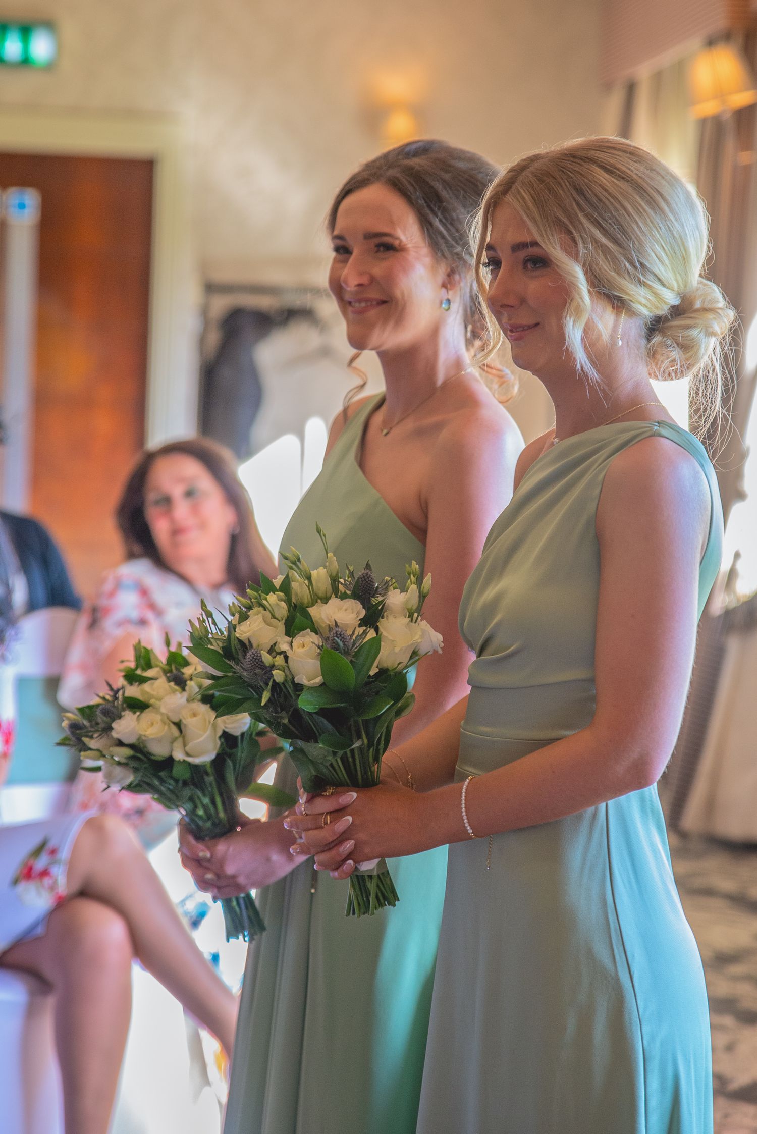 Bridesmaids in mint green dresses holding white rose bouquets at indoor venue.