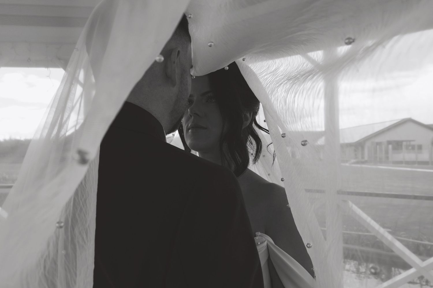 Black and white photo of a couple sharing an intimate moment under a flowing white veil outdoors.
