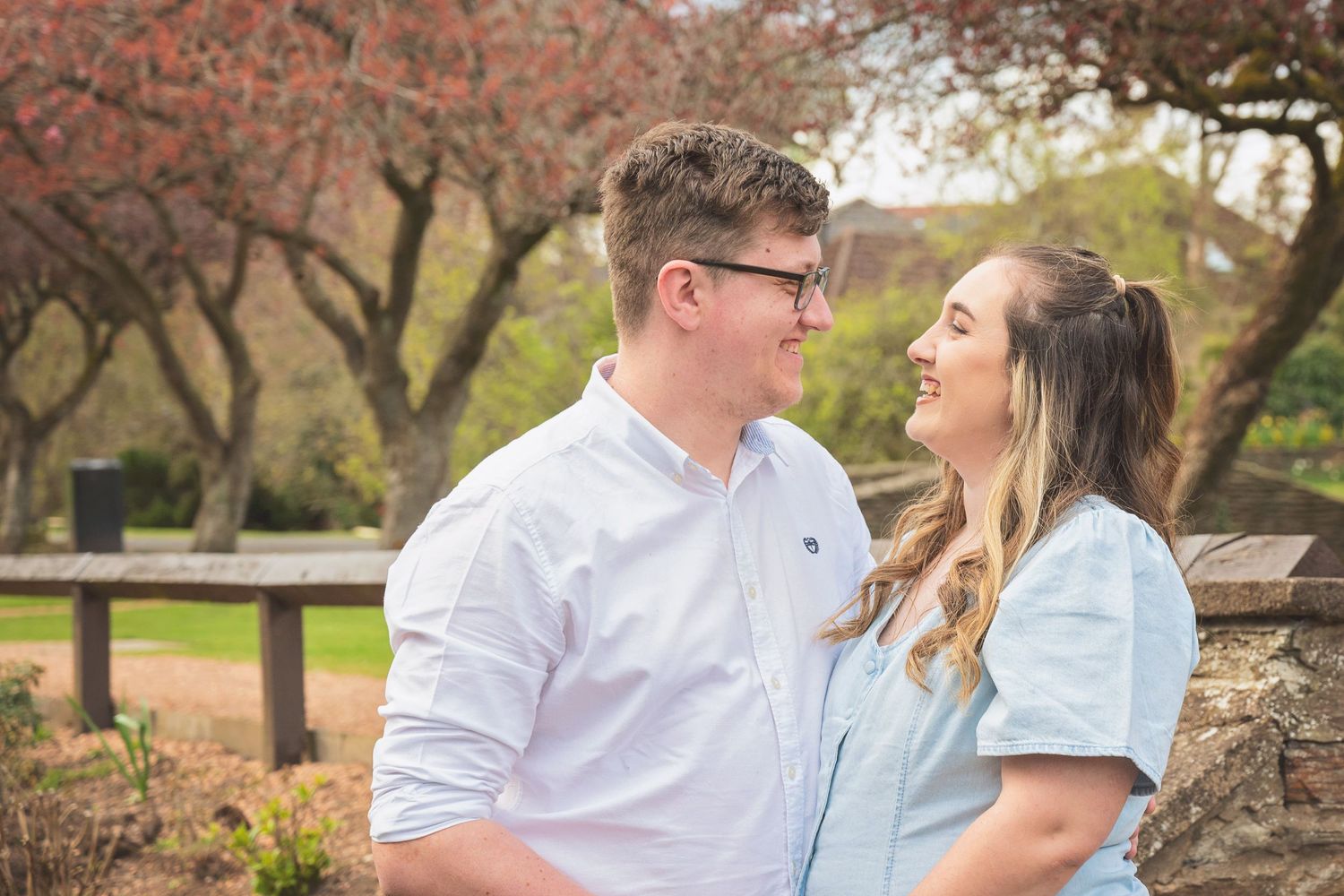 A smiling couple gazes at each other outdoors near a stone wall with pink flowering trees in the background.