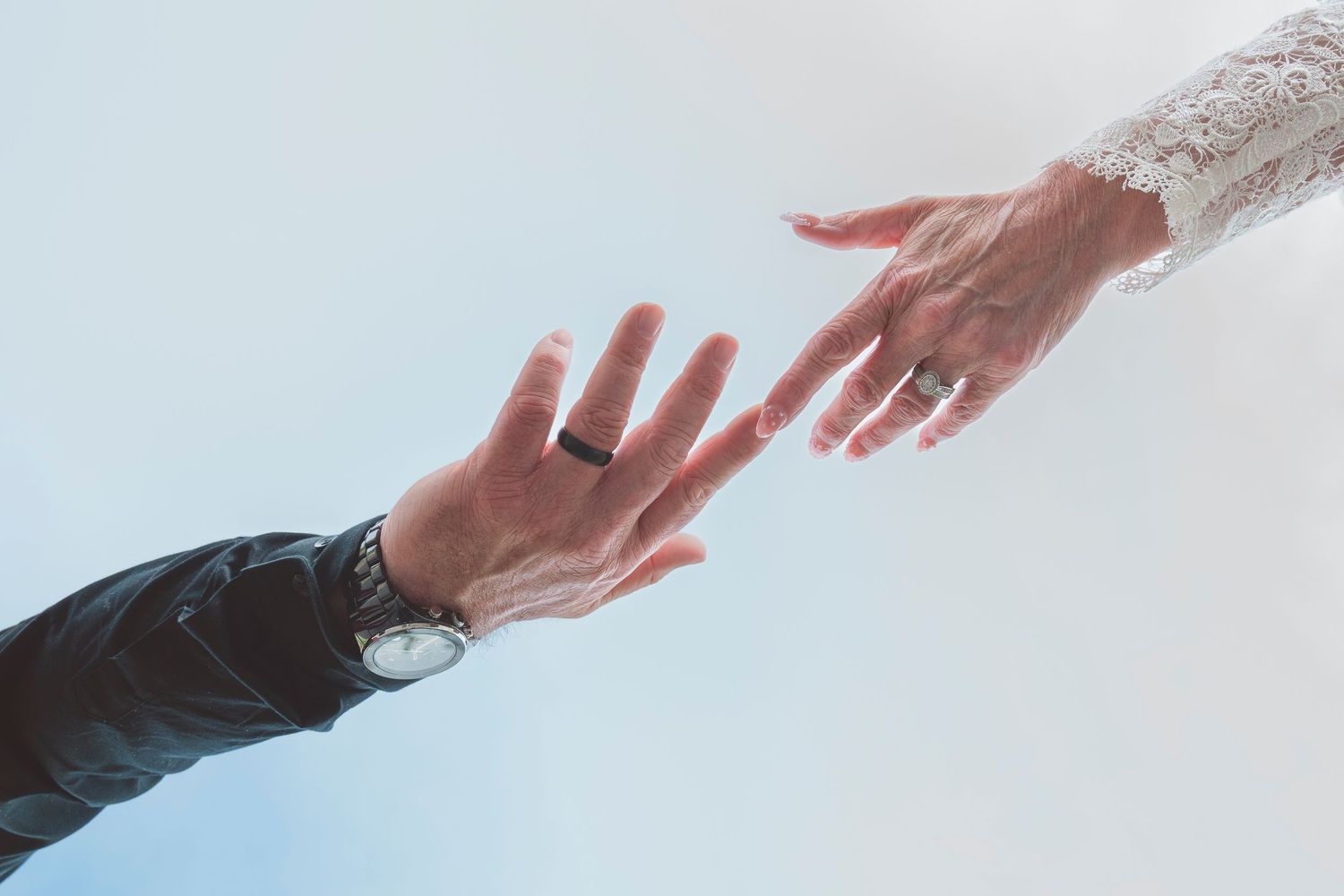 Two hands reaching toward each other, one wearing a wedding ring and watch, the other in lace, symbolizing marriage.