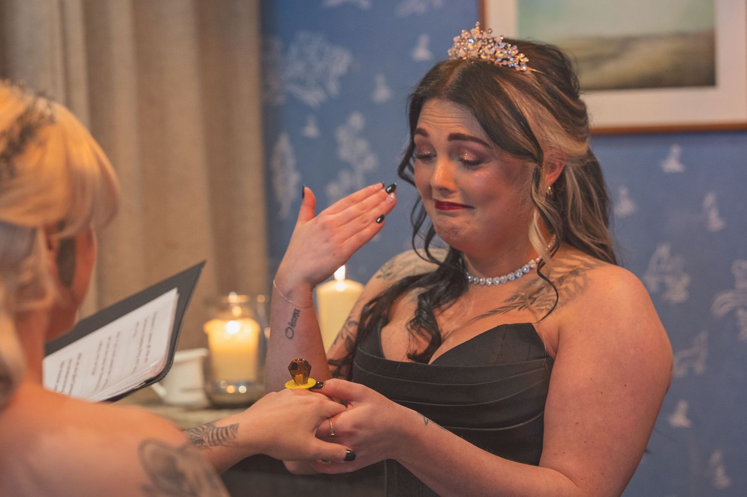 Emotional bride in black gown with tiara and diamond necklace reading a heartfelt note during wedding preparations.